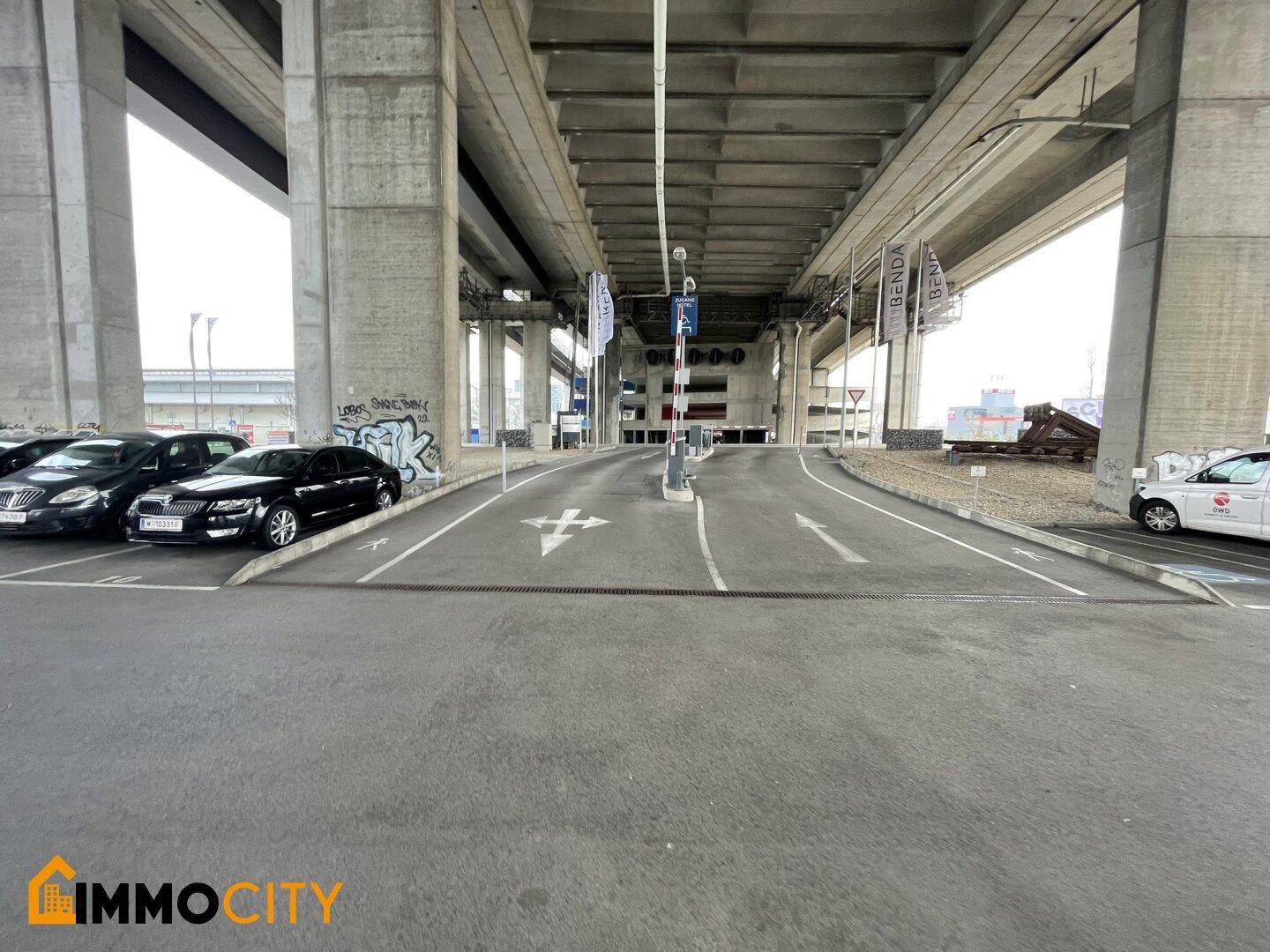 Einfahrt zu einem Parkhaus oder Parkbereich unter einer Autobahnbrücke mit klaren Fahrbahnmarkierungen.
