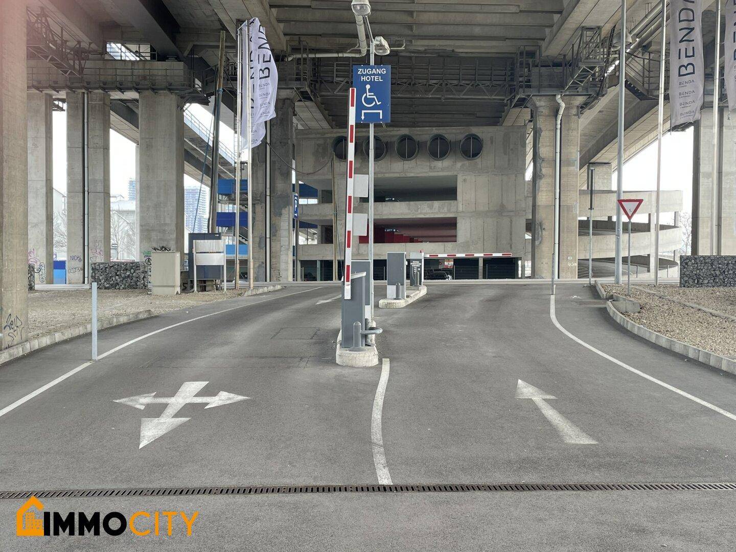 Eingangsbereich zu einem Parkplatz unter einer Brücke mit Schranke und Hinweisschildern für Hotelzugang.