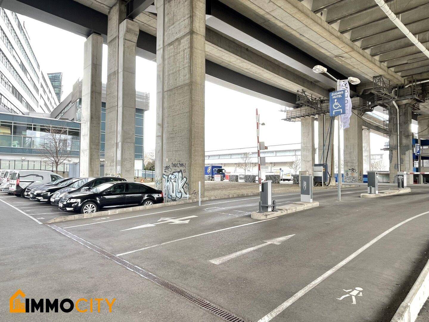 Zufahrt zu einem Parkbereich unter einer großen Betonbrücke, gekennzeichnet mit Pfeilen und Schranken.