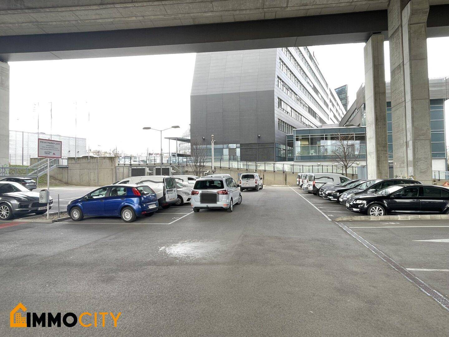 Blick auf einen Außenparkplatz unter einer Brücke mit geparkten Autos und einem modernen Gebäude im Hintergrund.