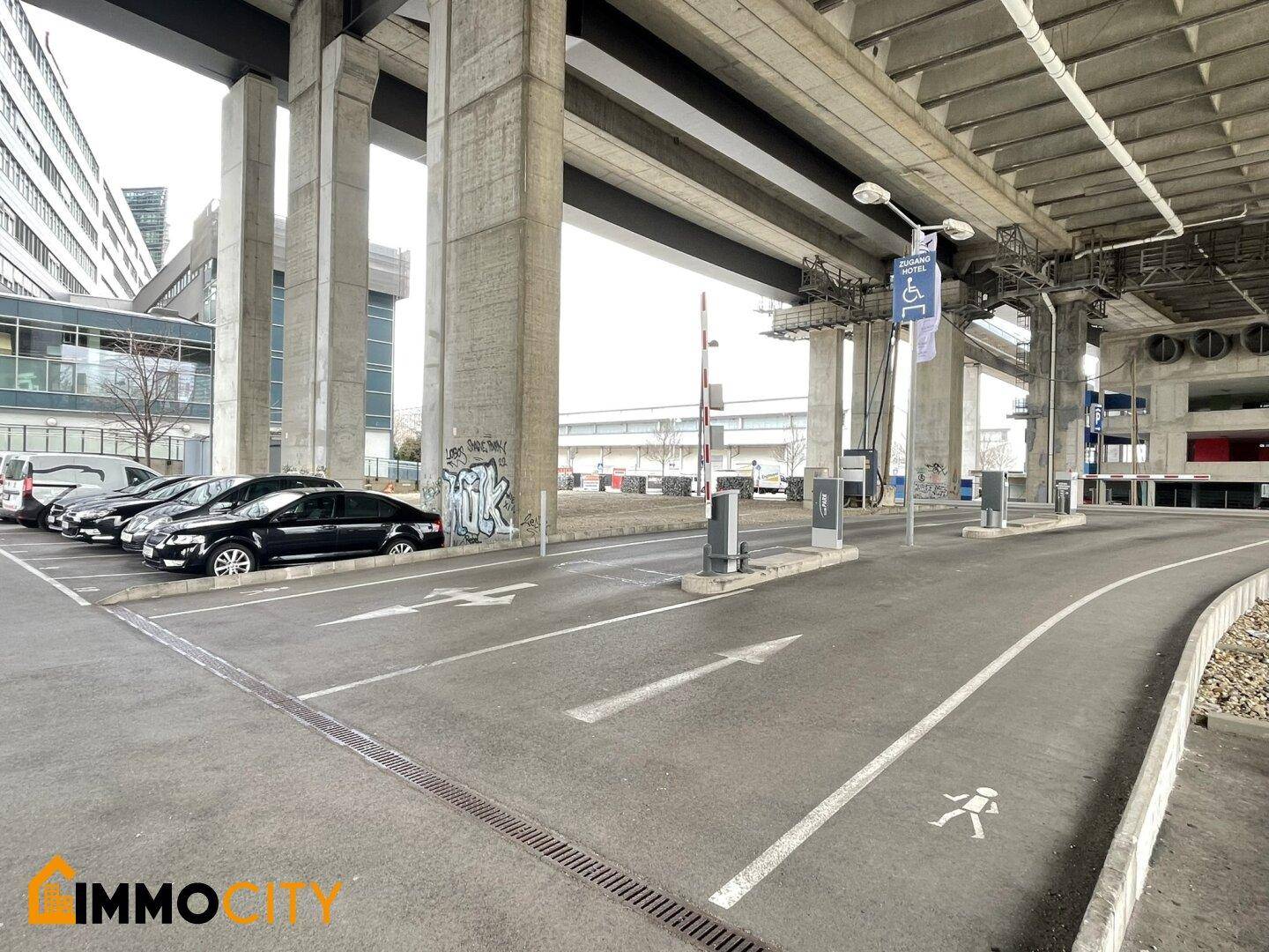 Zufahrt zu einem Parkbereich unter einer massiven Betonbrückenkonstruktion mit Fahrbahnmarkierungen und Schranken.