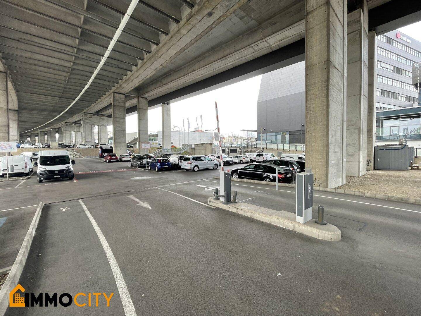 Weitläufiger Parkplatz unter einer Brücke mit zahlreichen Fahrzeugen und einer Zufahrtskontrolle im Vordergrund.
