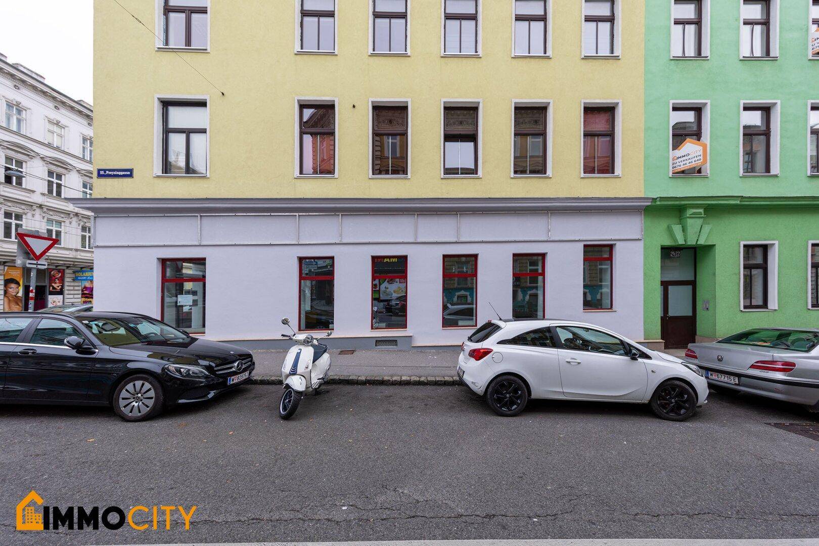 Straßenansicht des gelben Gebäudes mit angrenzendem grünem Gebäude und mehreren geparkten Fahrzeugen.
