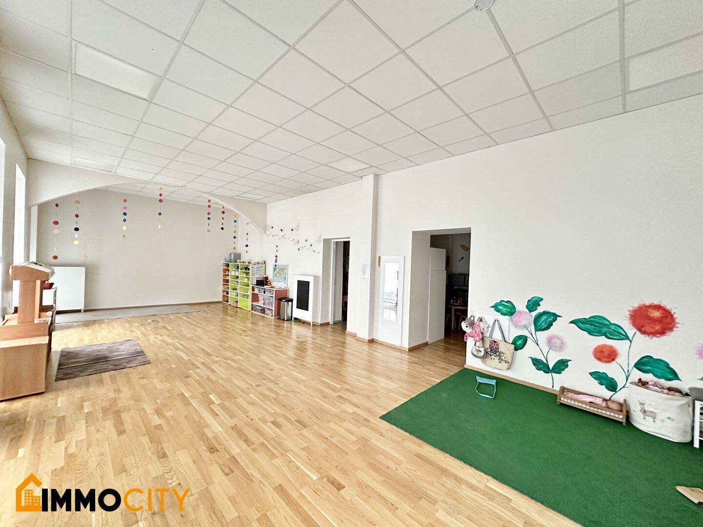 Großer, heller Spielraum mit Holzboden, bunten Wanddekorationen und Regalen für Spielzeug.