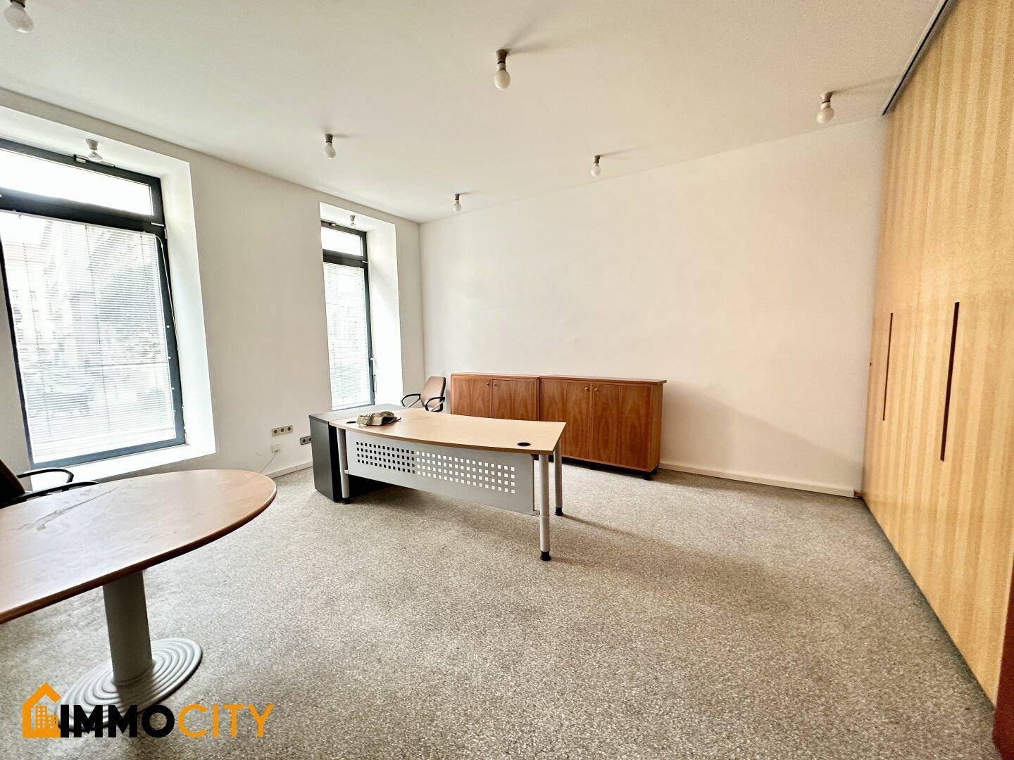 Helles Büro mit Teppichboden, modernen Schreibtischen und zwei großen Fenstern zur Straße.