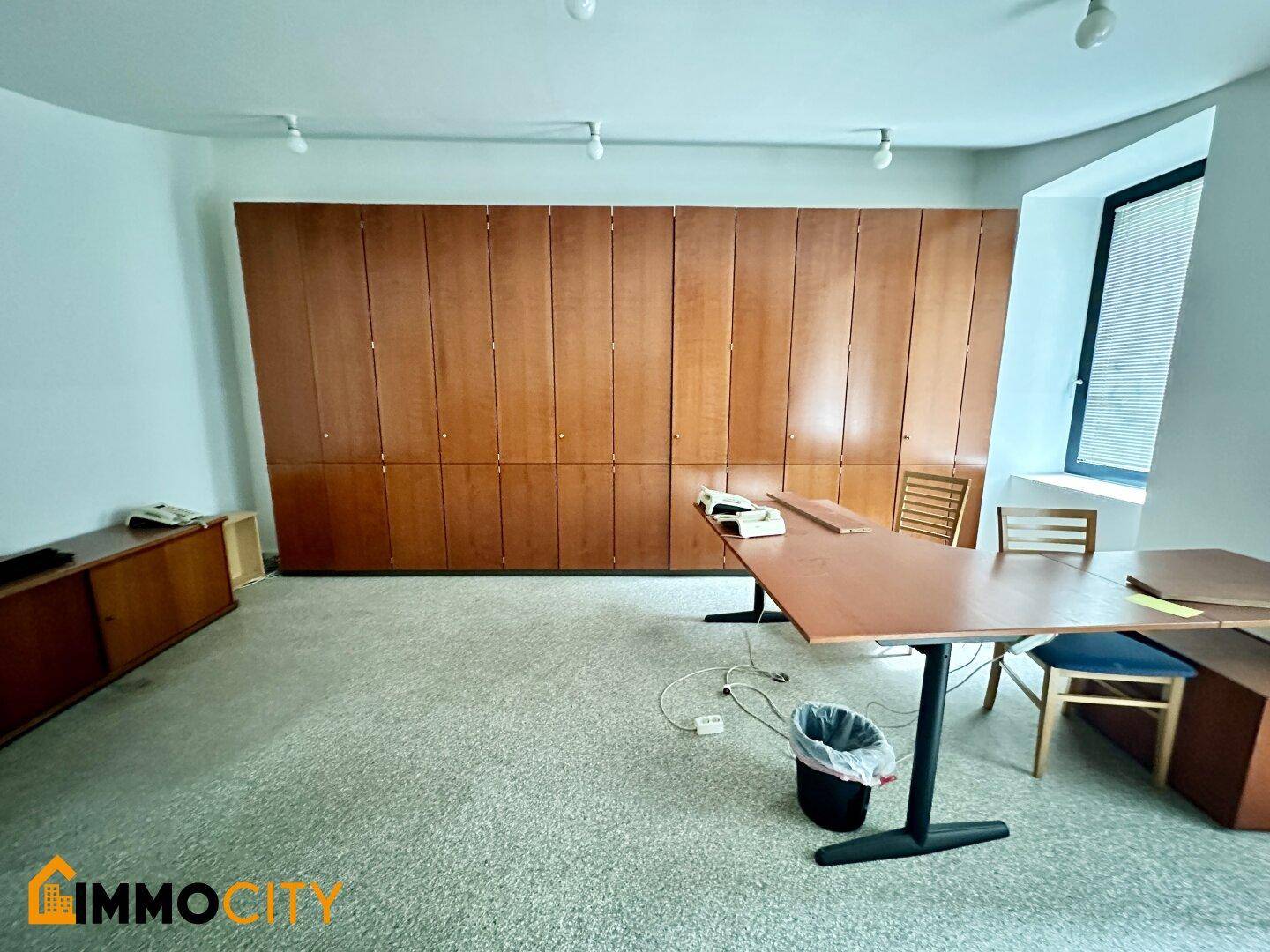 Büro mit umfangreichen Holz-Einbauschränken, Teppichboden und einem Fenster mit Jalousien.