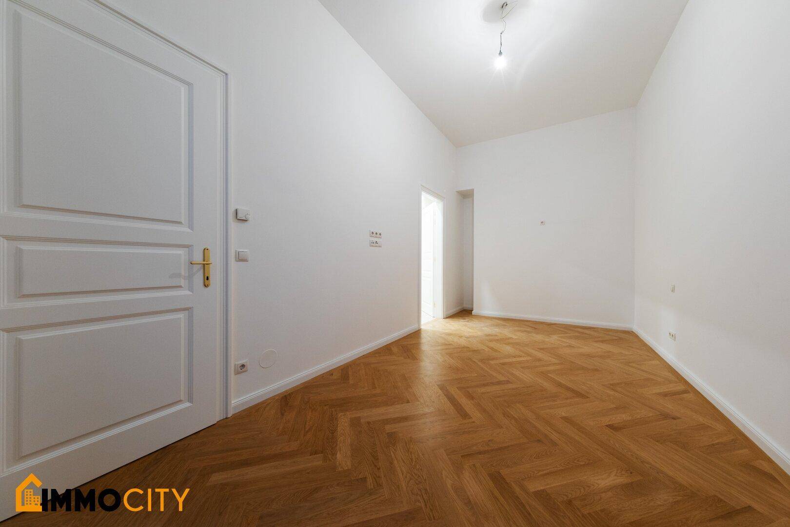 Helles Zimmer mit Fischgrätparkett und einer weißen Tür, bereit für individuelle Einrichtungsideen.