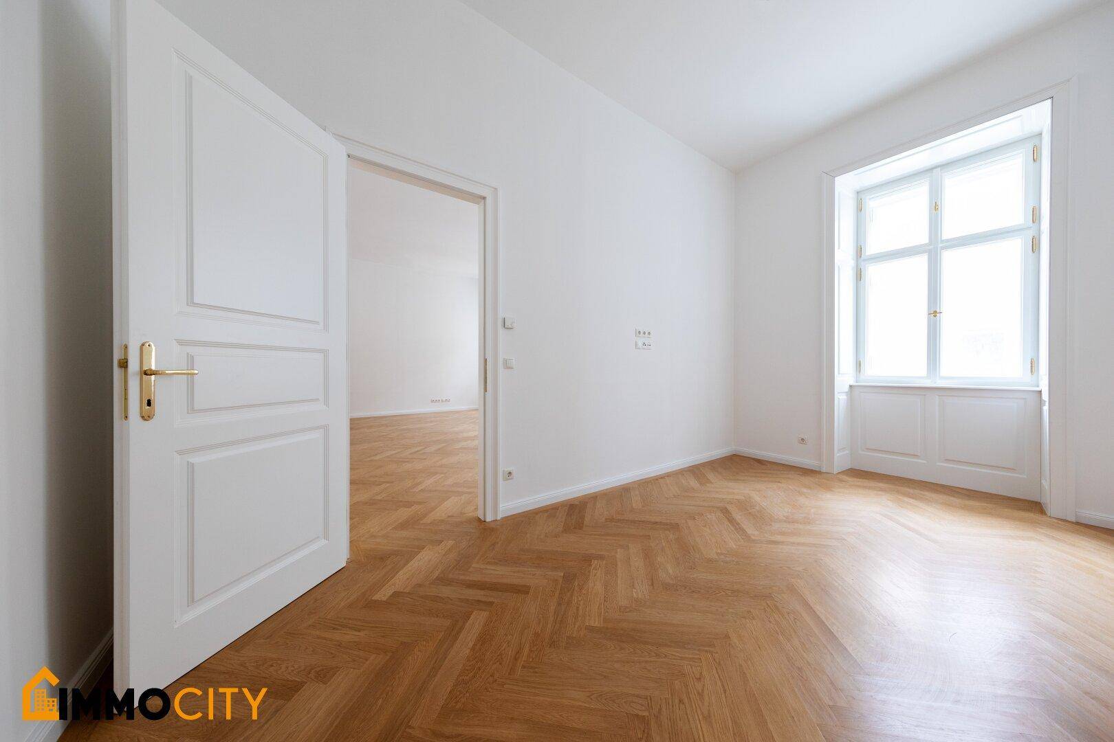 Geräumiges Zimmer mit Fischgrätparkett und einem großen Fenster, das viel Tageslicht hereinlässt.