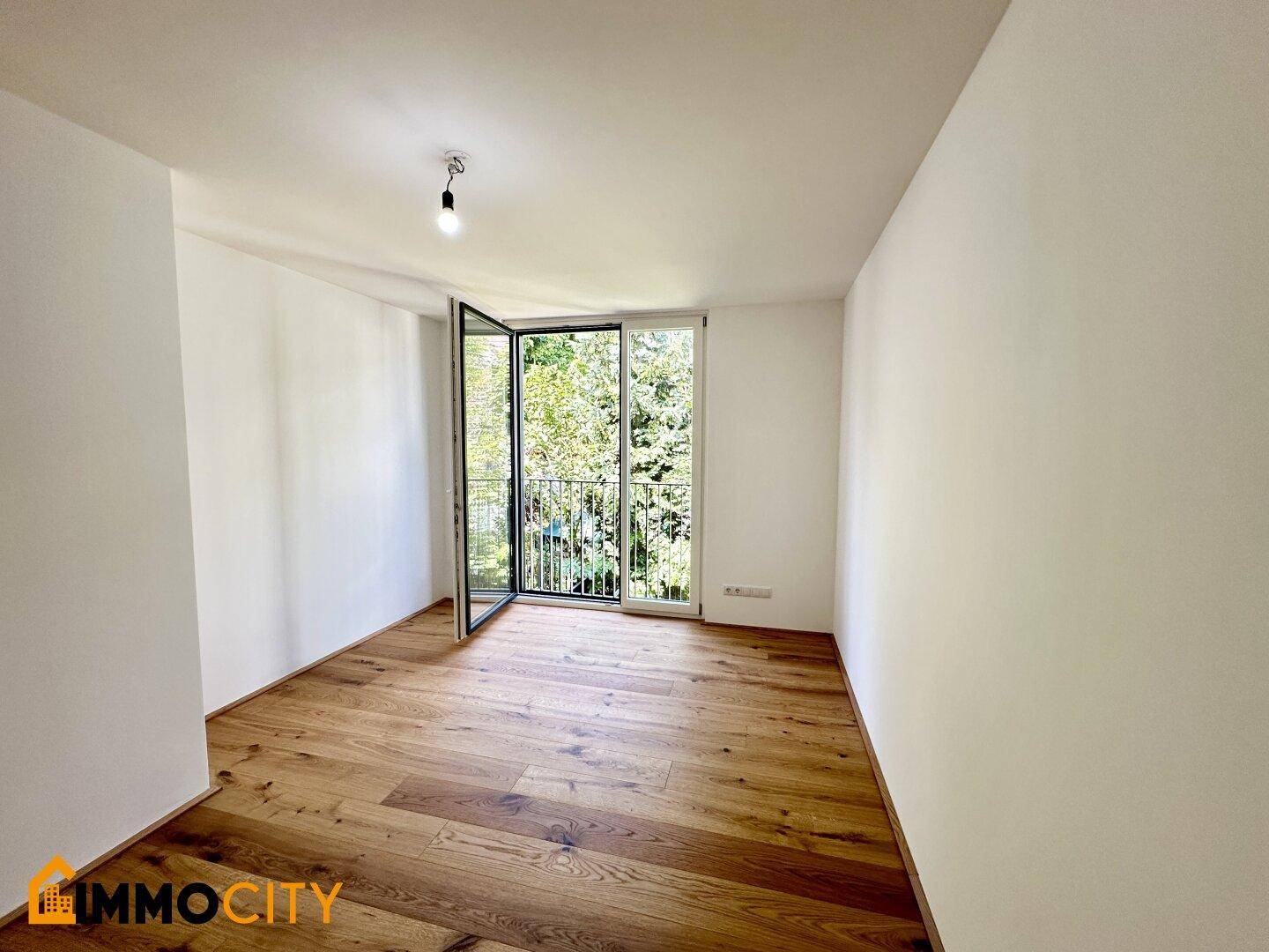Helles Zimmer mit Eichenparkettboden und Zugang zu einem französischen Balkon mit Grünblick.