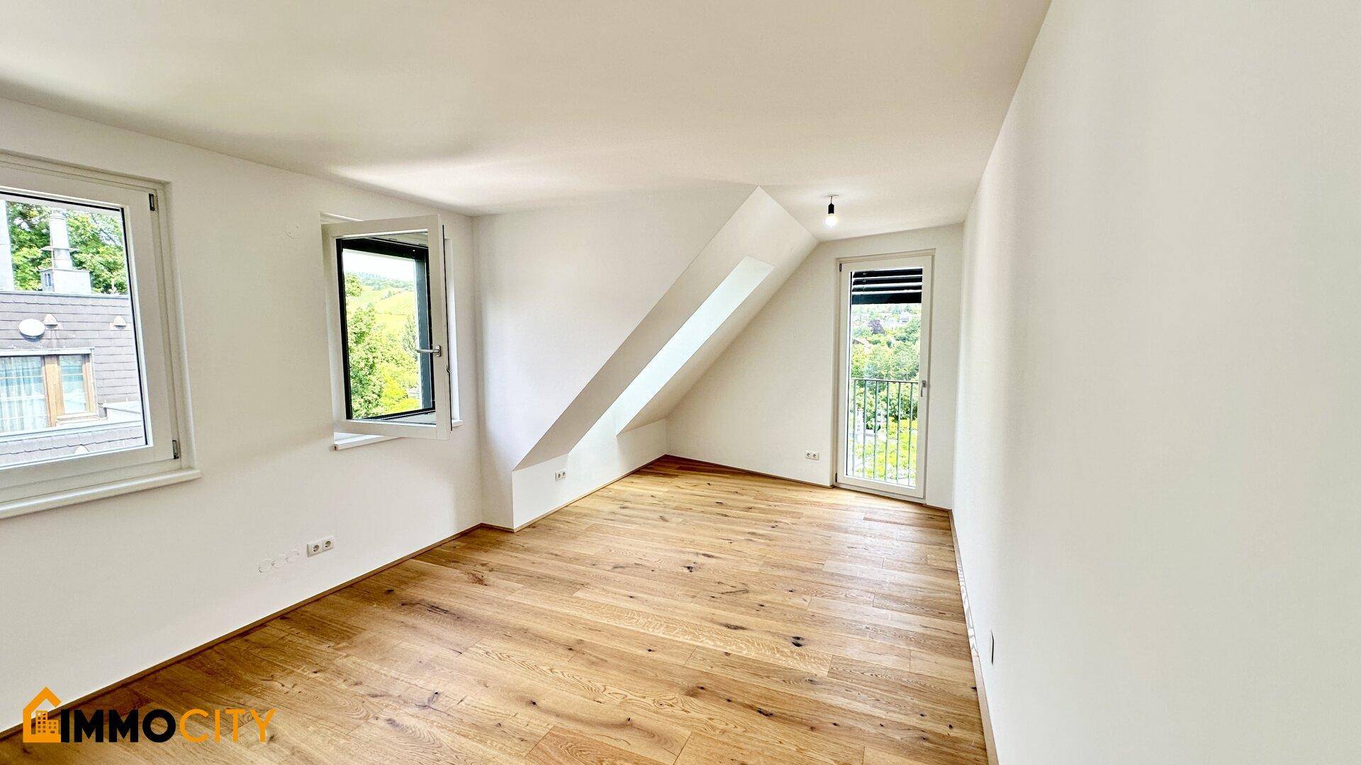 Geräumiges Zimmer mit Holzboden, schräger Decke und Fenstern mit Blick ins Grüne.