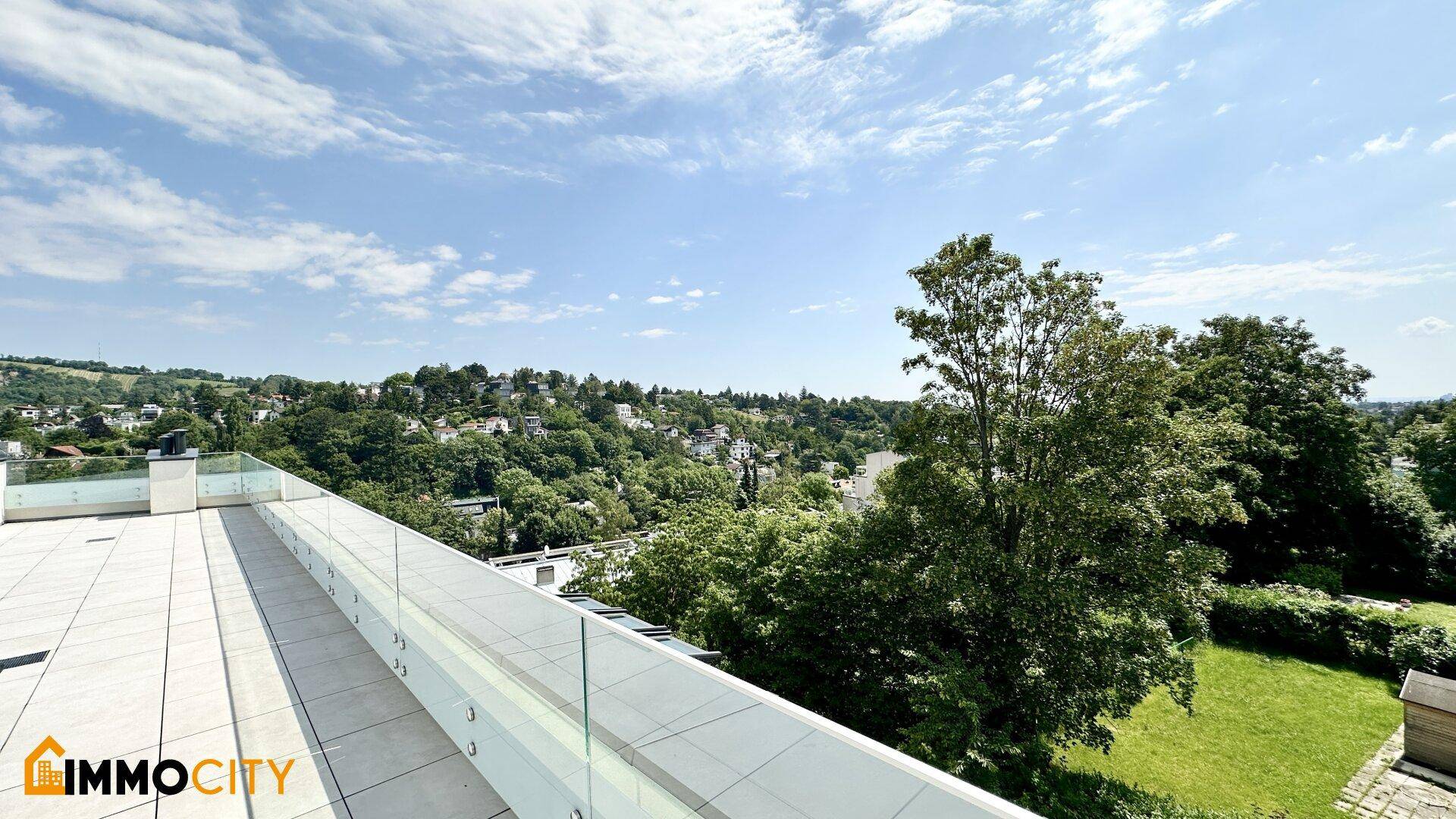 Helle Dachterrasse mit Glasbrüstung und Blick auf eine grüne Wiese sowie die umliegende Wohngegend.