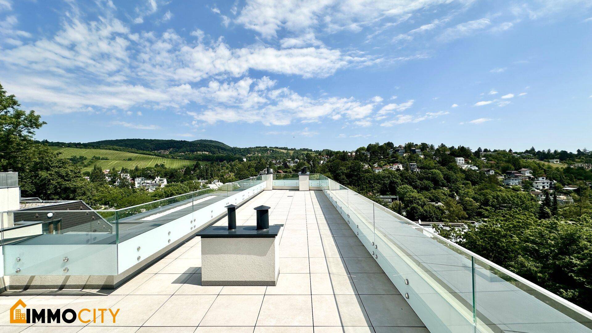 Weitläufige Dachterrasse mit Fliesenboden, Glasgeländer und Panoramablick über die bewaldete Umgebung.