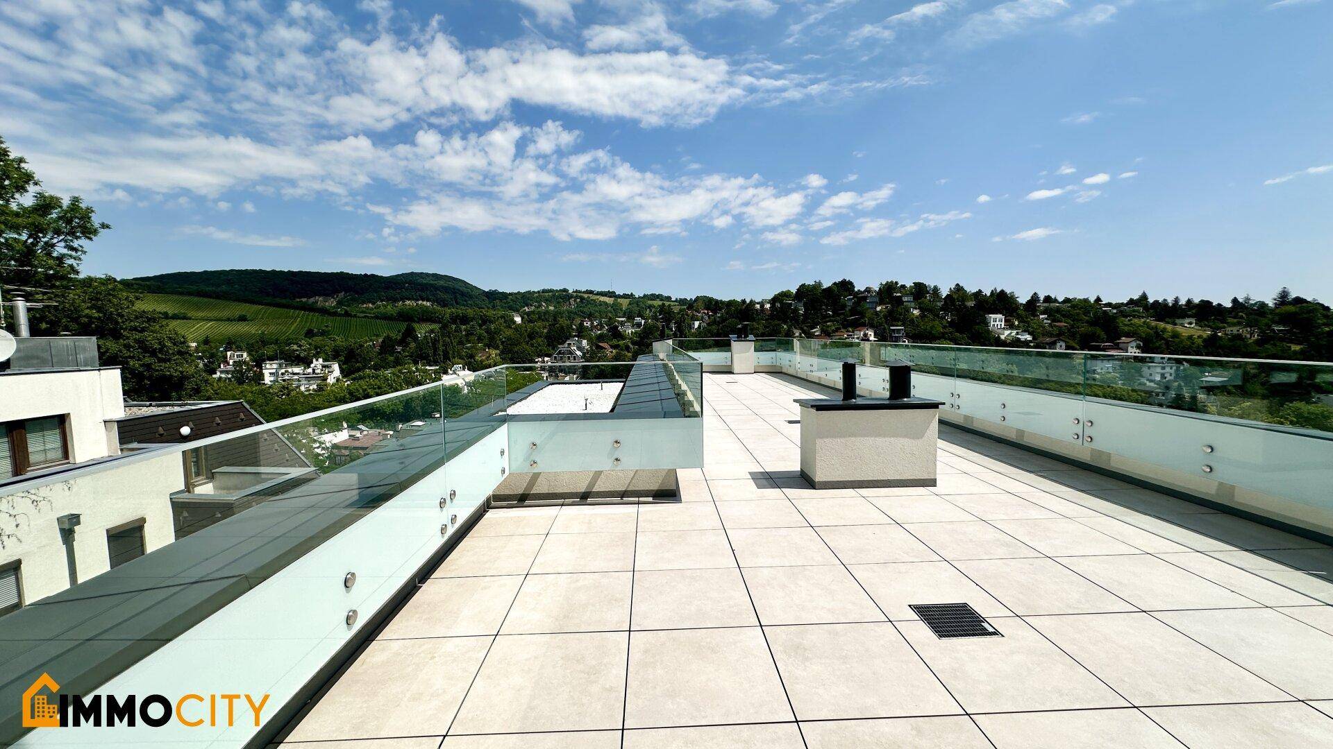 Weitläufige Dachterrasse mit Fliesenboden, Glasgeländer und Panoramablick über die bewaldete Umgebung.