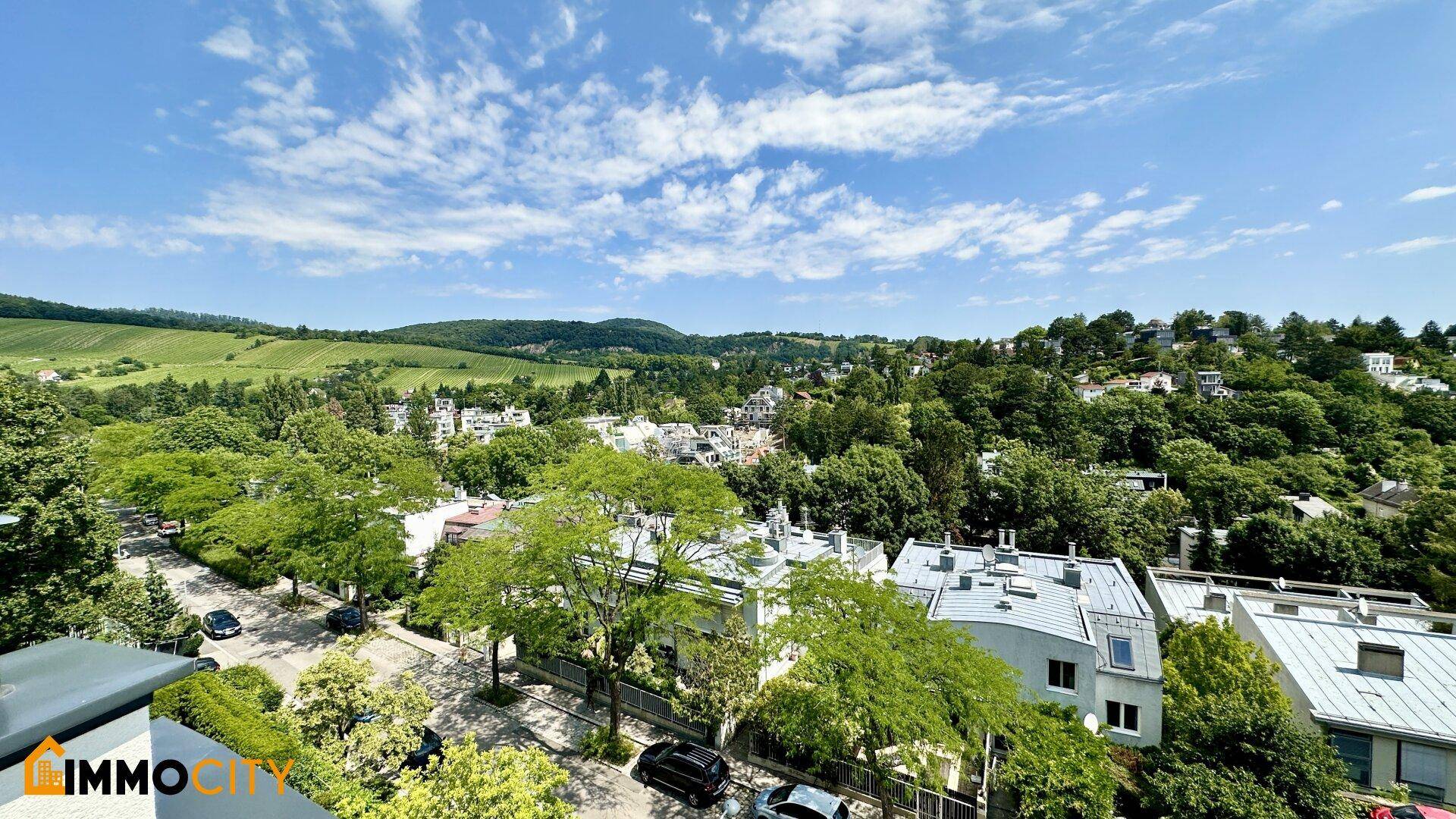 Weitläufiger Blick über grüne Hügel, bewaldete Gebiete und eine Wohnsiedlung unter blauem Himmel.