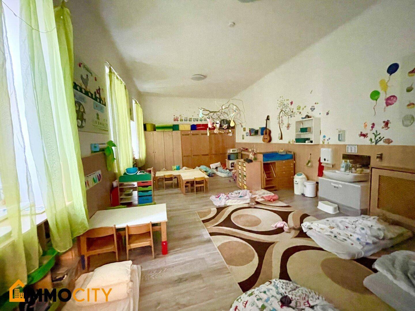 Gemütlicher Ruhebereich für Kinder mit Matratzen, kleinen Tischen und hellen Fenstern mit grünen Vorhängen.