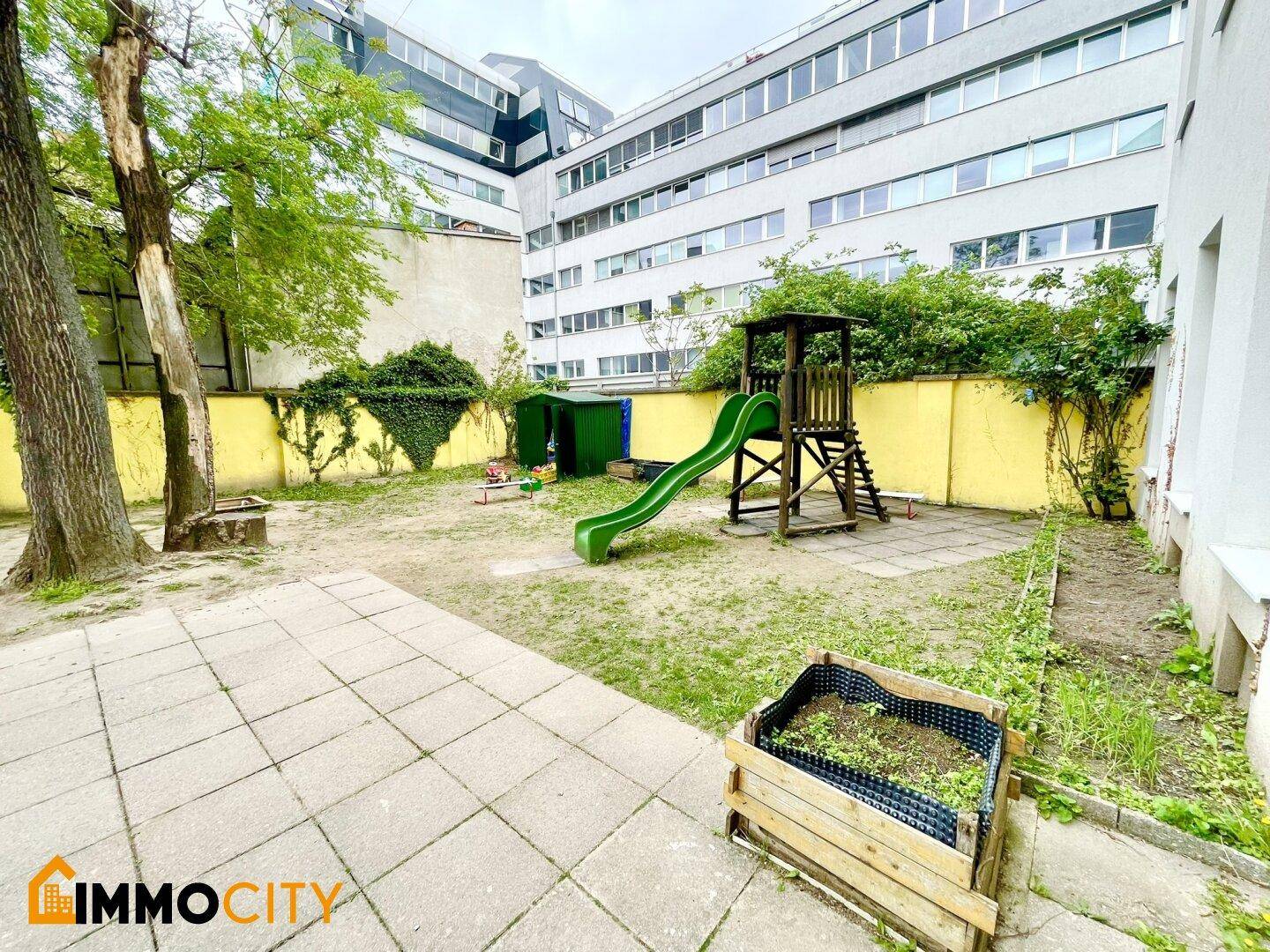 Außenbereich mit Holzspielturm, Rutsche und Hochbeet, umgeben von Bäumen und Gebäuden.