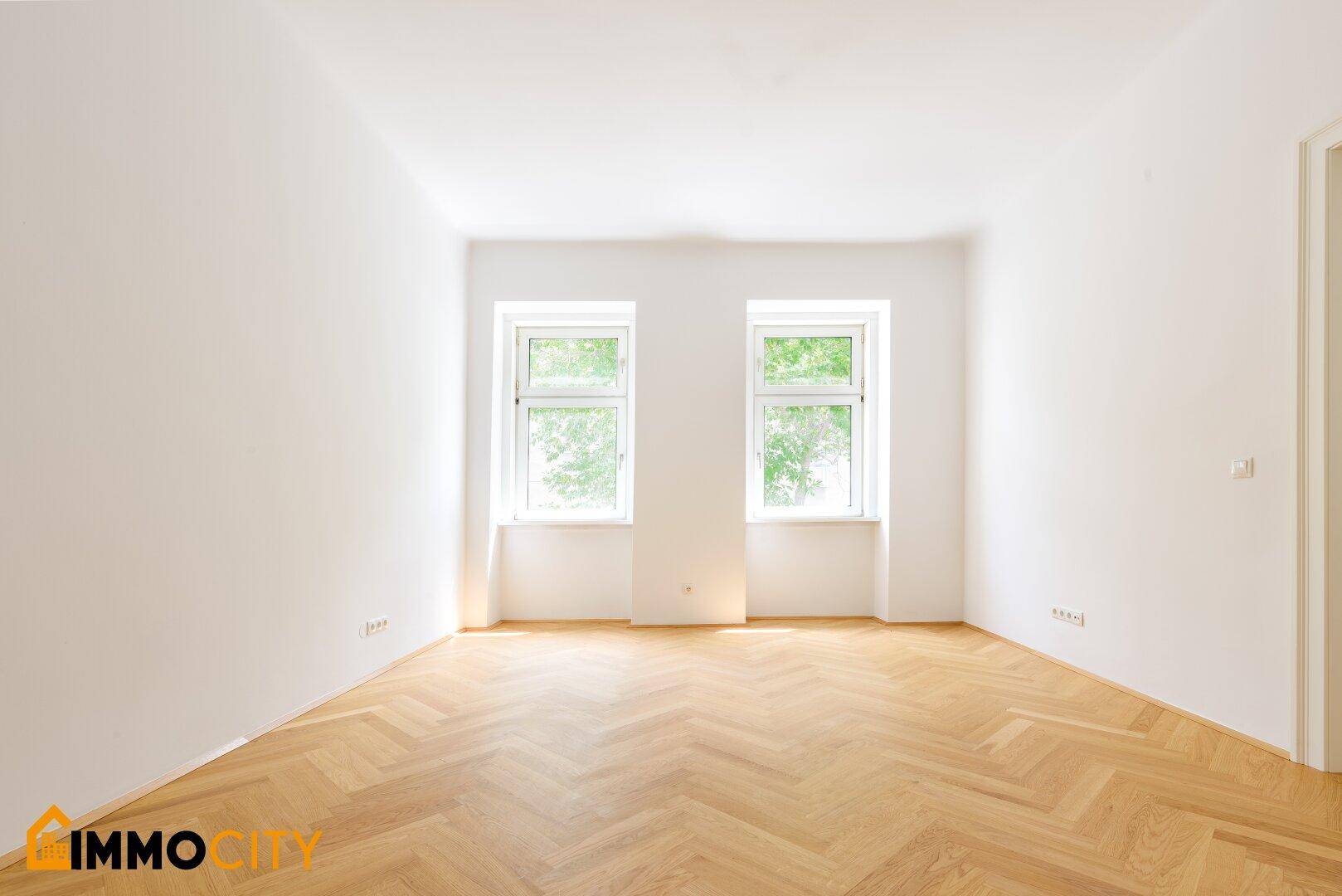 Geräumiges Zimmer mit hellem Fischgrätparkett, weißen Wänden und zwei großen Fenstern.