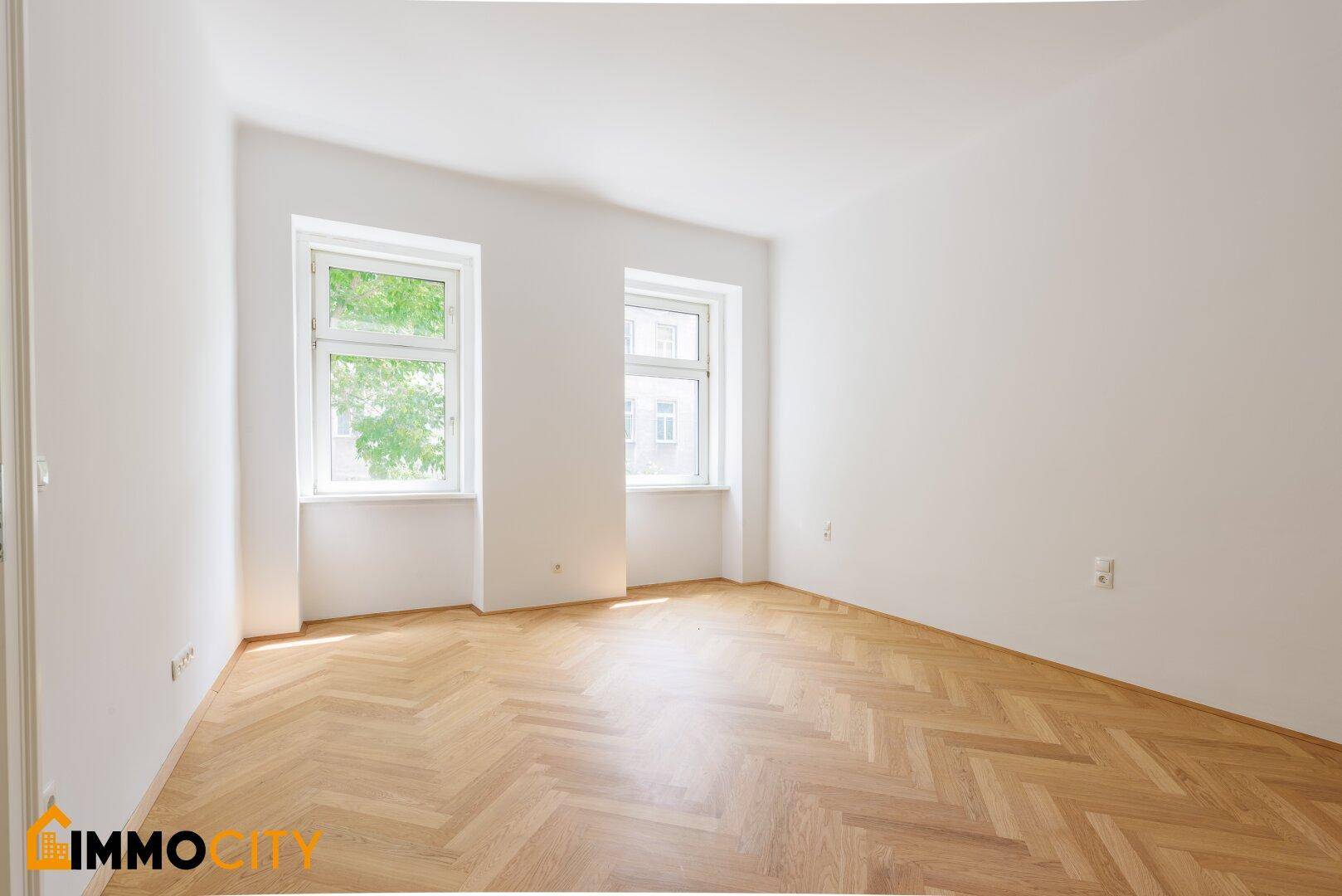 Lichtdurchflutetes Zimmer mit klassischem Fischgrätparkett und Blick ins Grüne durch die Fenster.