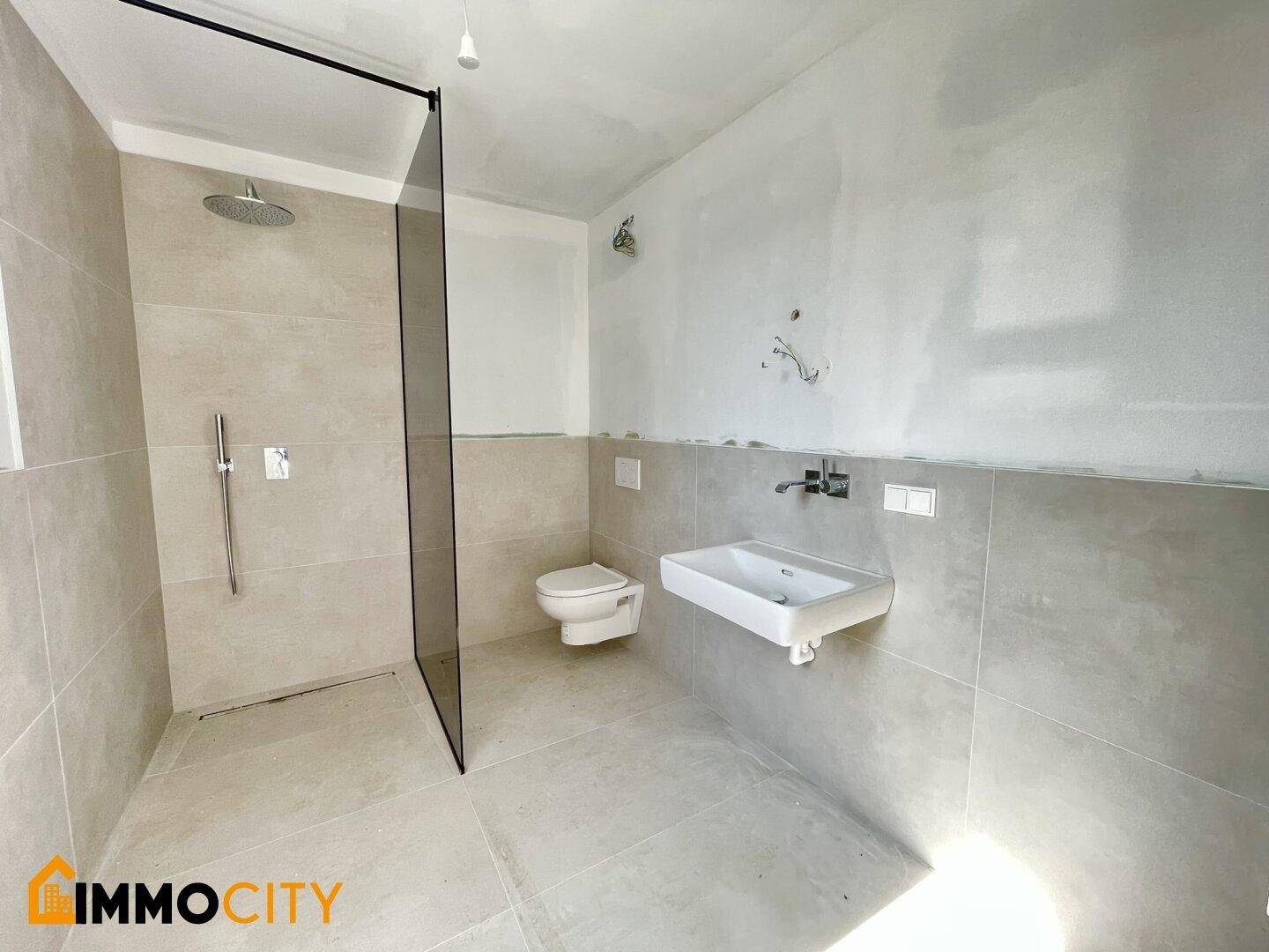 Modernes Badezimmer im Rohbau mit Fliesenboden, wandhängendem WC und begehbarer Dusche.