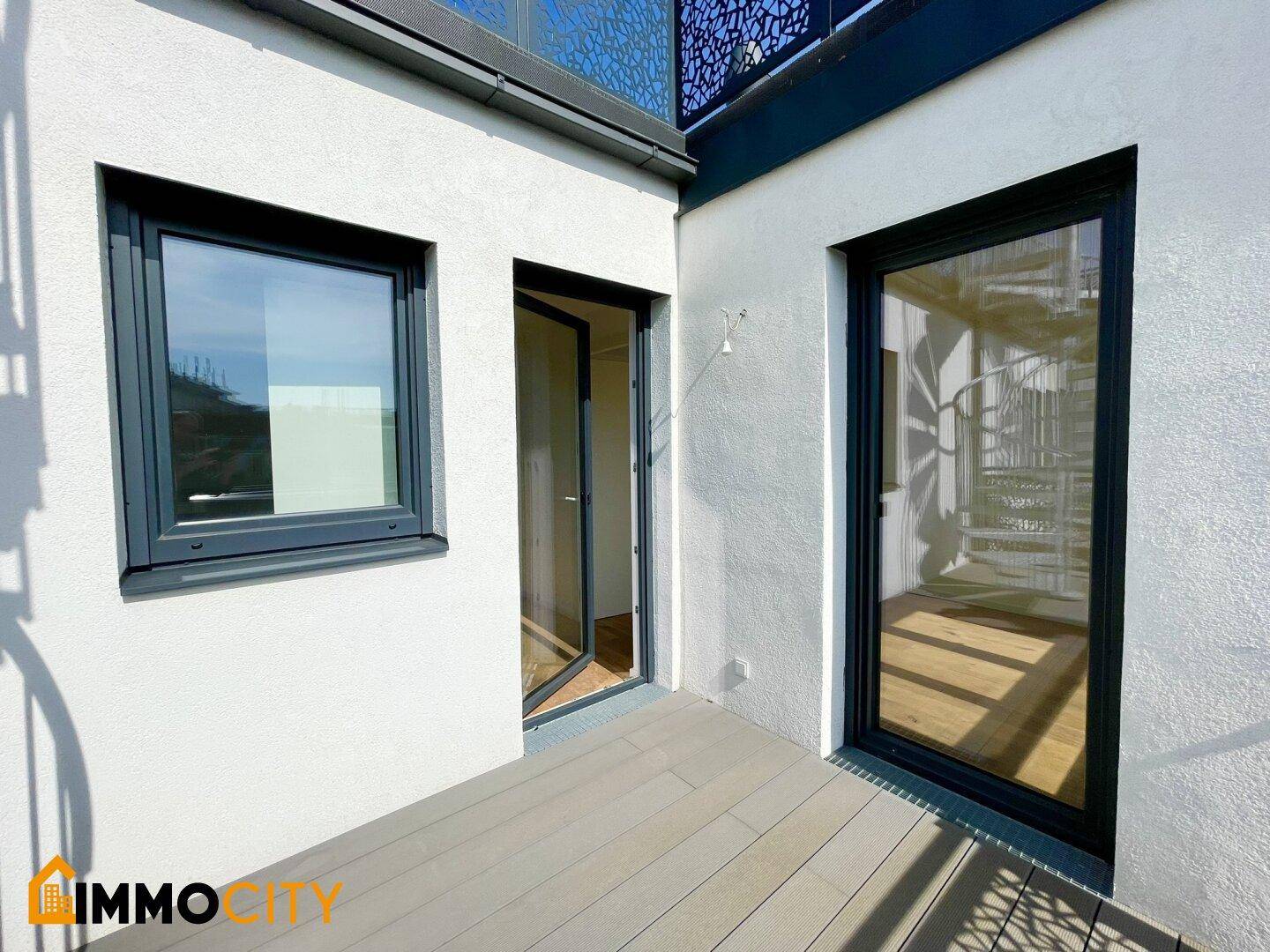 Außenbereich mit Holzboden, Fenster und zwei Türen, die zu einem sonnigen Außenbereich führen.