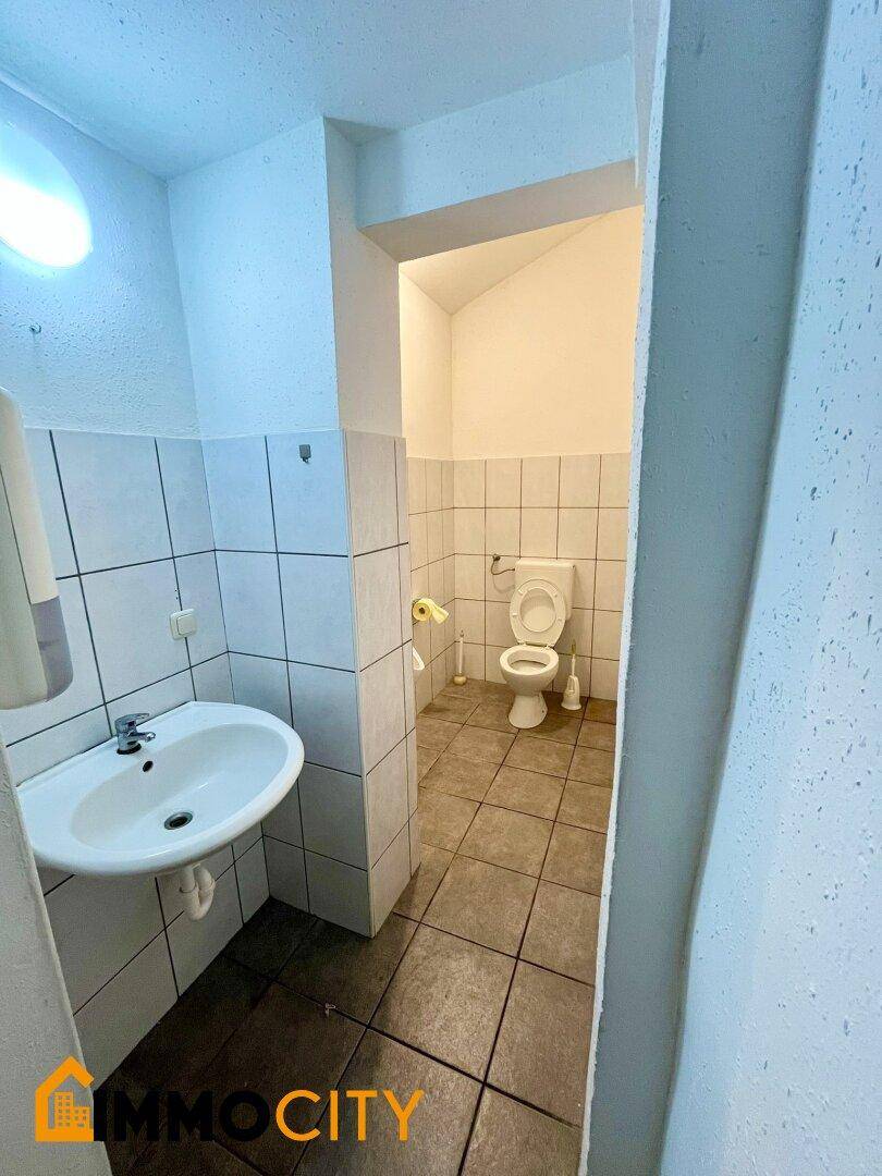 Toilettenbereich mit WC und Waschbecken, umgeben von weißen Wandfliesen.