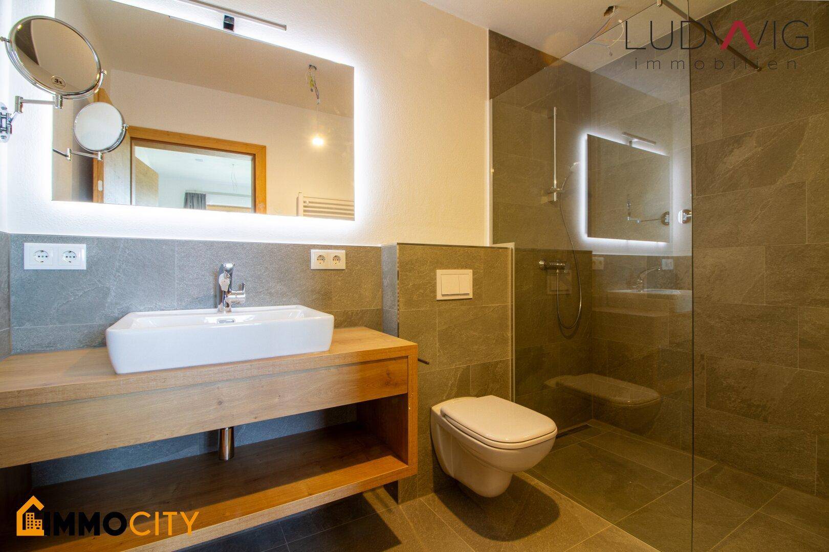 Stilvolles Badezimmer mit großem beleuchtetem Spiegel, Waschtisch und begehbarer Dusche.