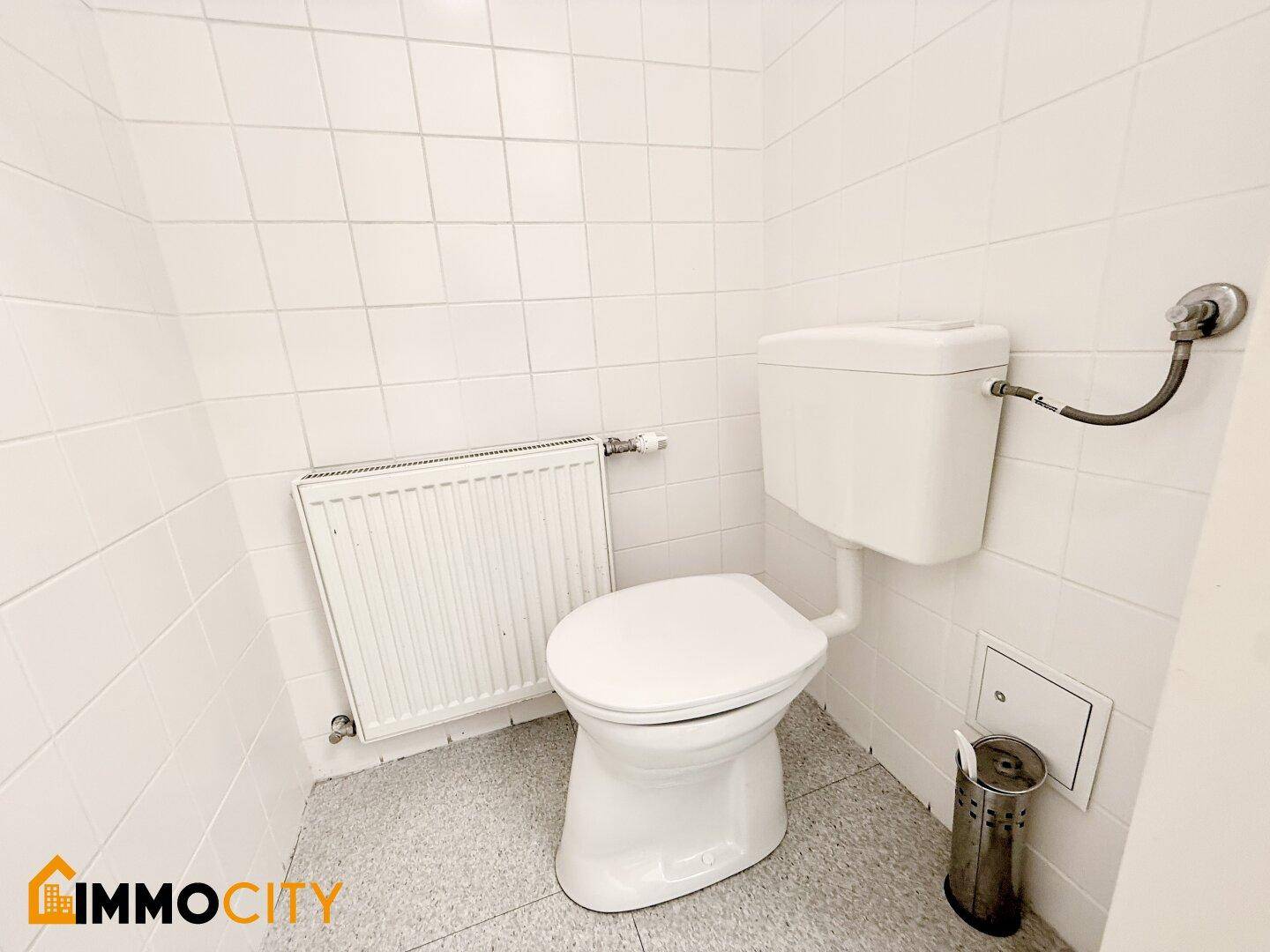 Einfache Toilette mit Spülkasten und Heizkörper, gefliesten Wänden und Boden.
