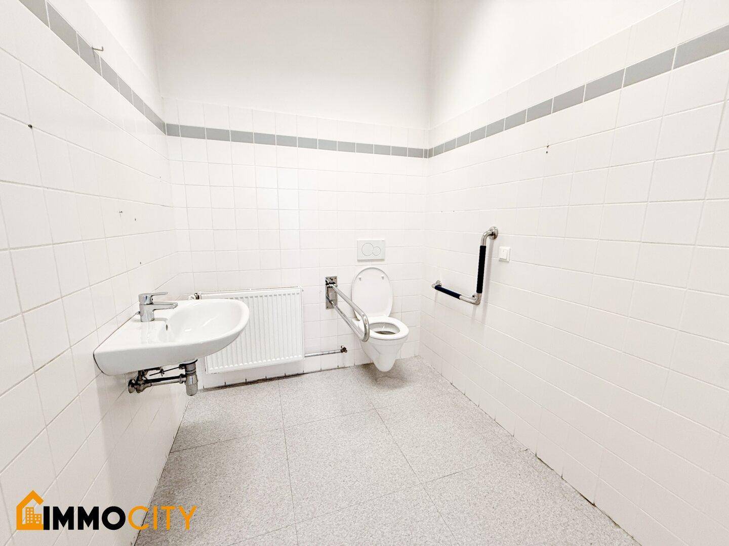Barrierefreies WC mit Haltegriffen, Waschbecken und Fliesen an Wänden und Boden.