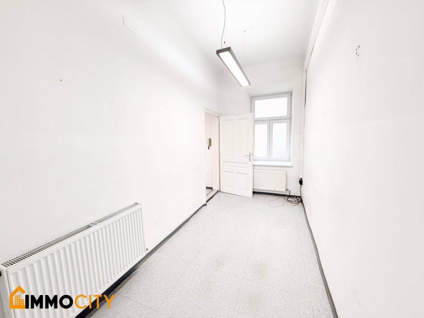 Langer, schmaler Büroraum mit Fenster, Heizkörper und einfacher Beleuchtung, bereit zur Nutzung.