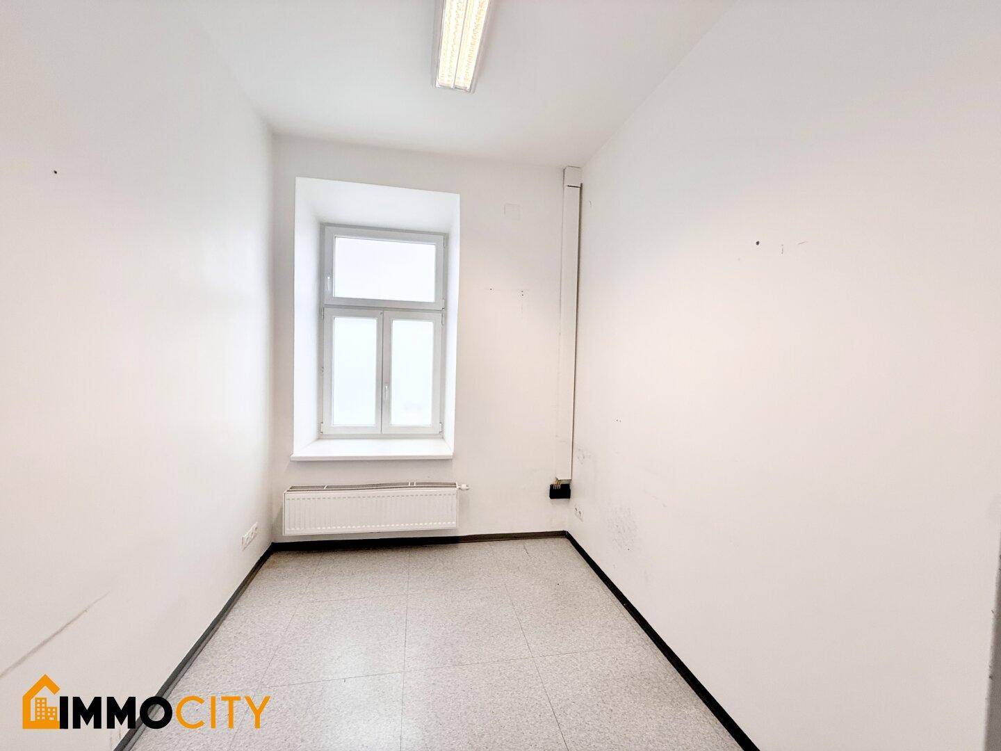 Kleiner Büroraum mit Fenster, Heizkörper und Fliesenboden, bietet eine einfache und funktionale Arbeitsumgebung.