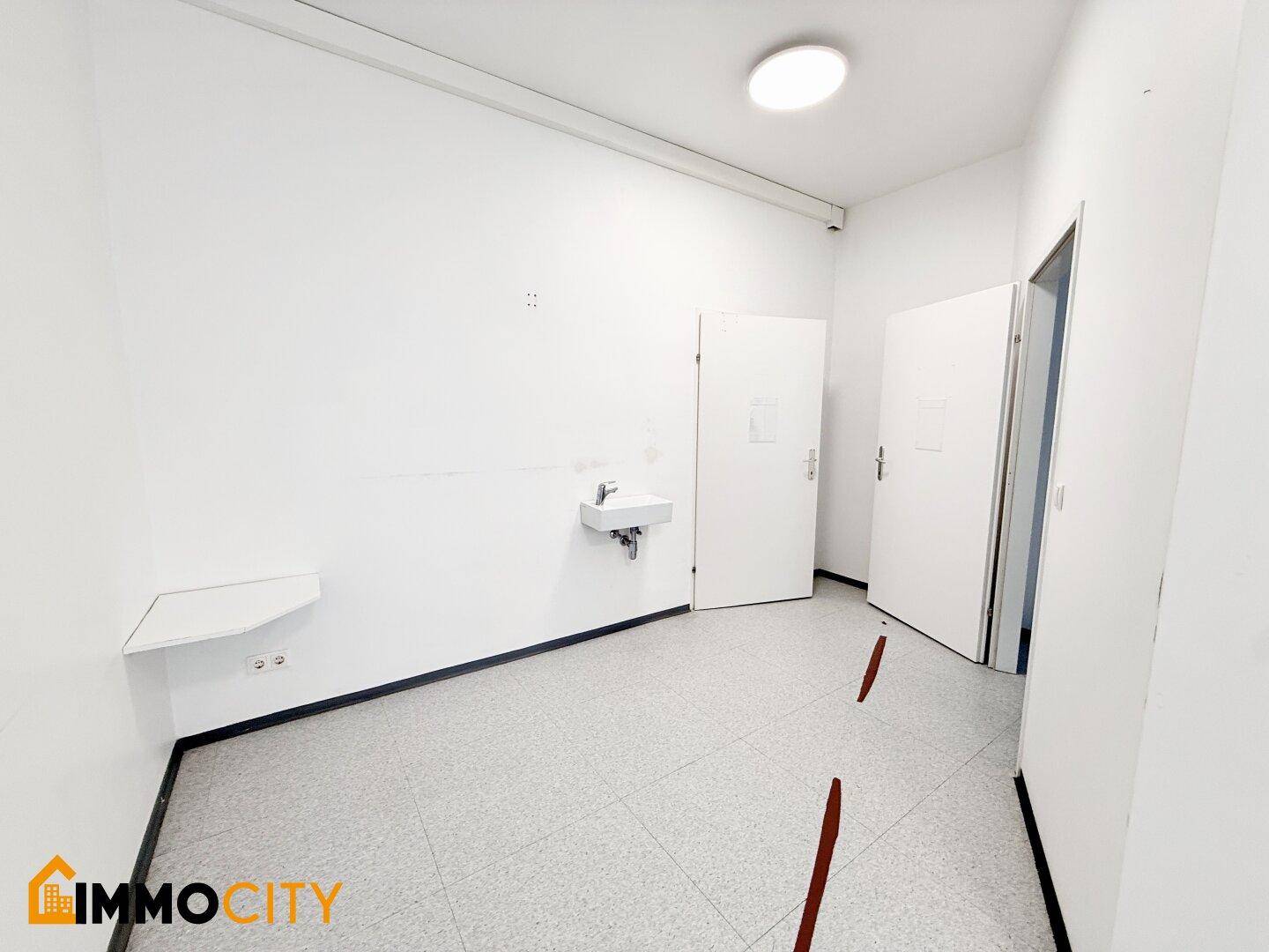 Heller Büroraum mit Waschbecken, zwei Türen und Fliesenboden, ausgestattet mit einfacher Beleuchtung.