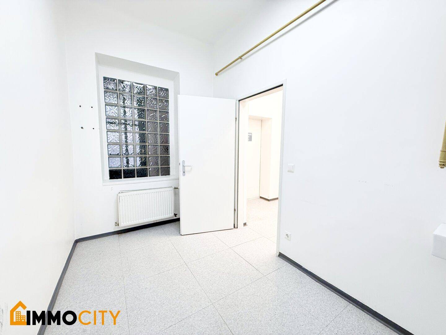 Kleiner Büroraum mit Fenster aus Glasbausteinen, Heizkörper und Fliesenboden, ideal für ein Einzelbüro.