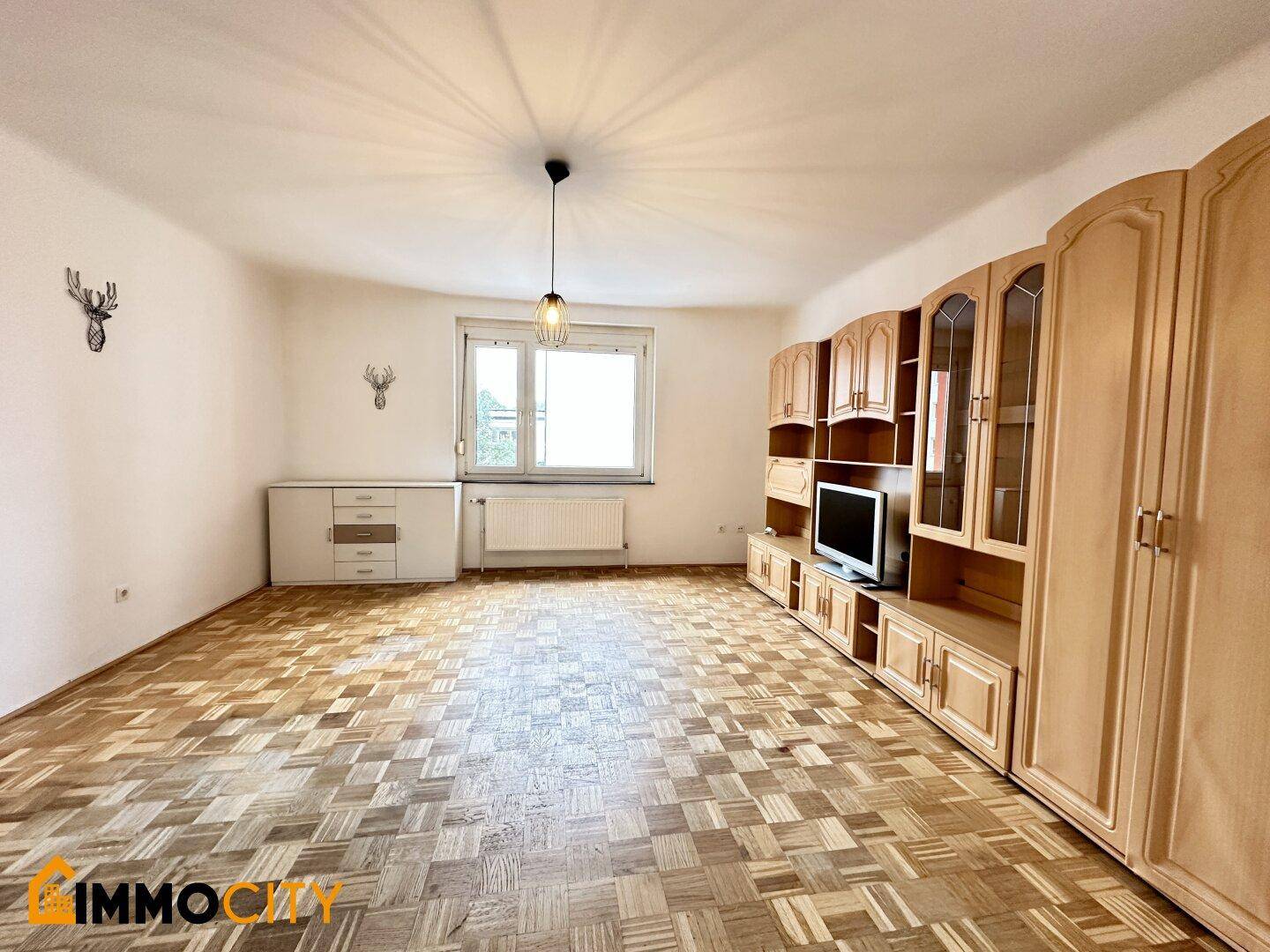 Helles Wohnzimmer mit Parkettboden, großem Fenster und einer geräumigen Schrankwand.