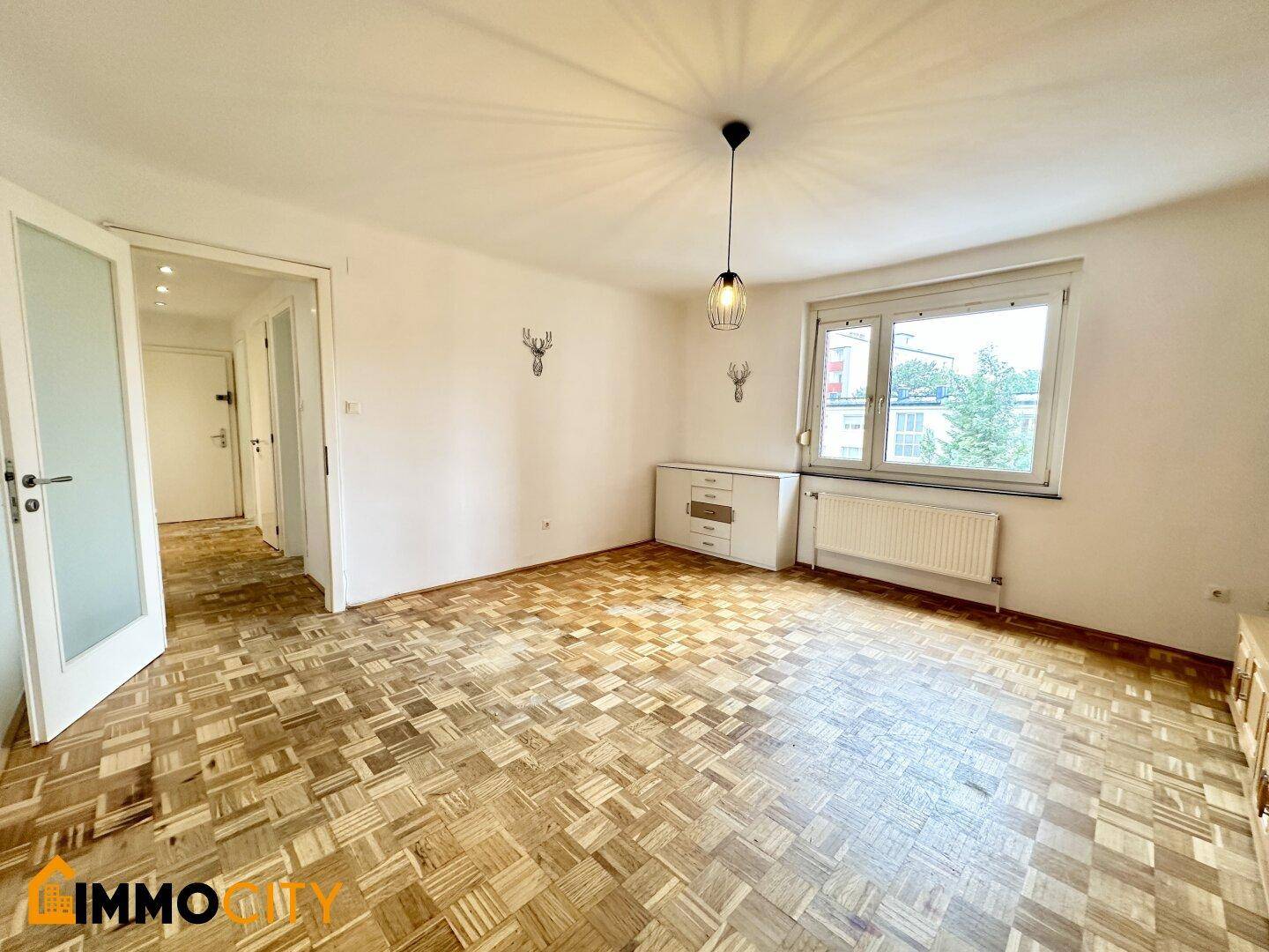 Helles Wohnzimmer mit Parkettboden, großem Fenster und Zugang zu weiteren Räumen.
