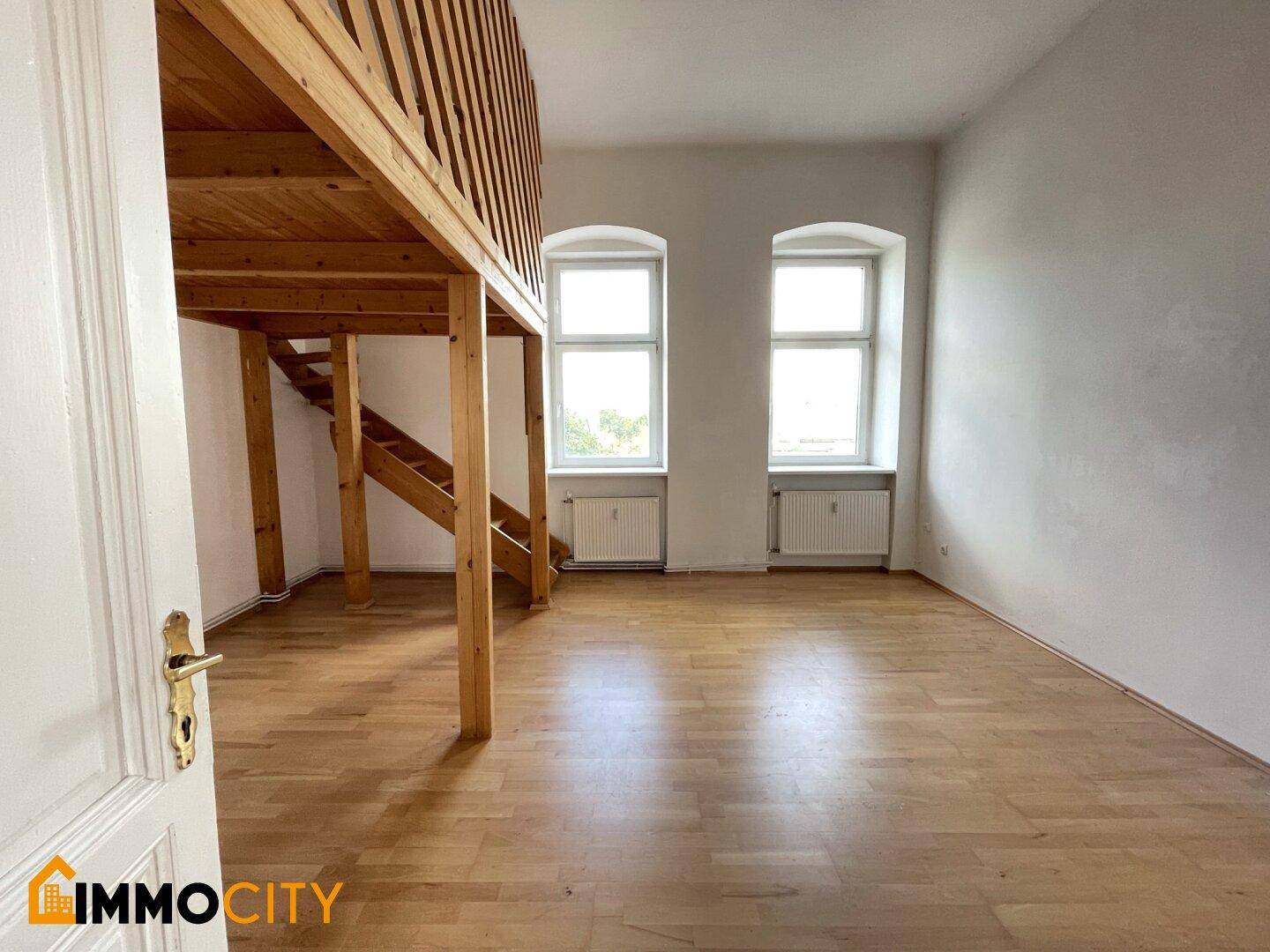 Geräumiger Wohnbereich mit Holzgalerie und zwei großen Fenstern, die viel Licht hereinlassen.