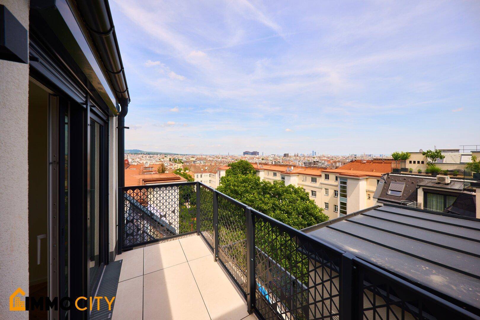 Geräumiger Balkon mit Metallgeländer und weitem Blick über die Stadt und grüne Bäume.