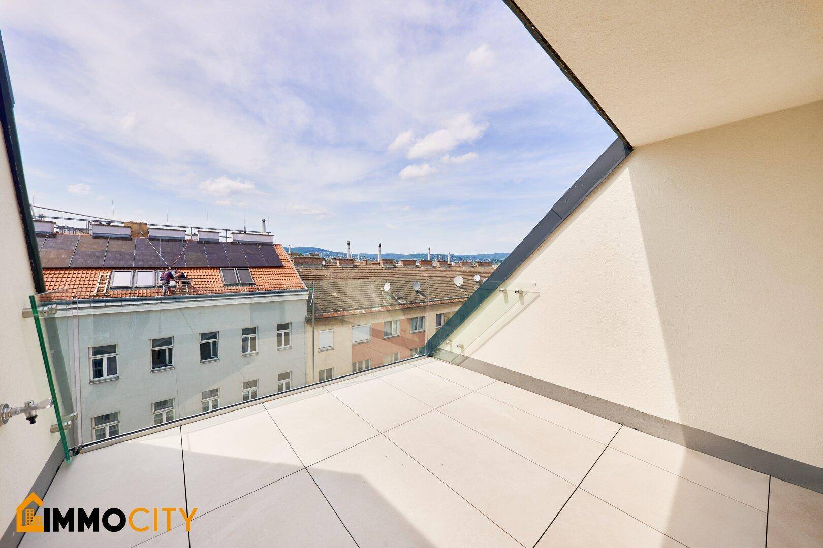 Kleiner Balkon mit Glasgeländer und Blick auf die Dächer der Stadt und die Berge im Hintergrund.