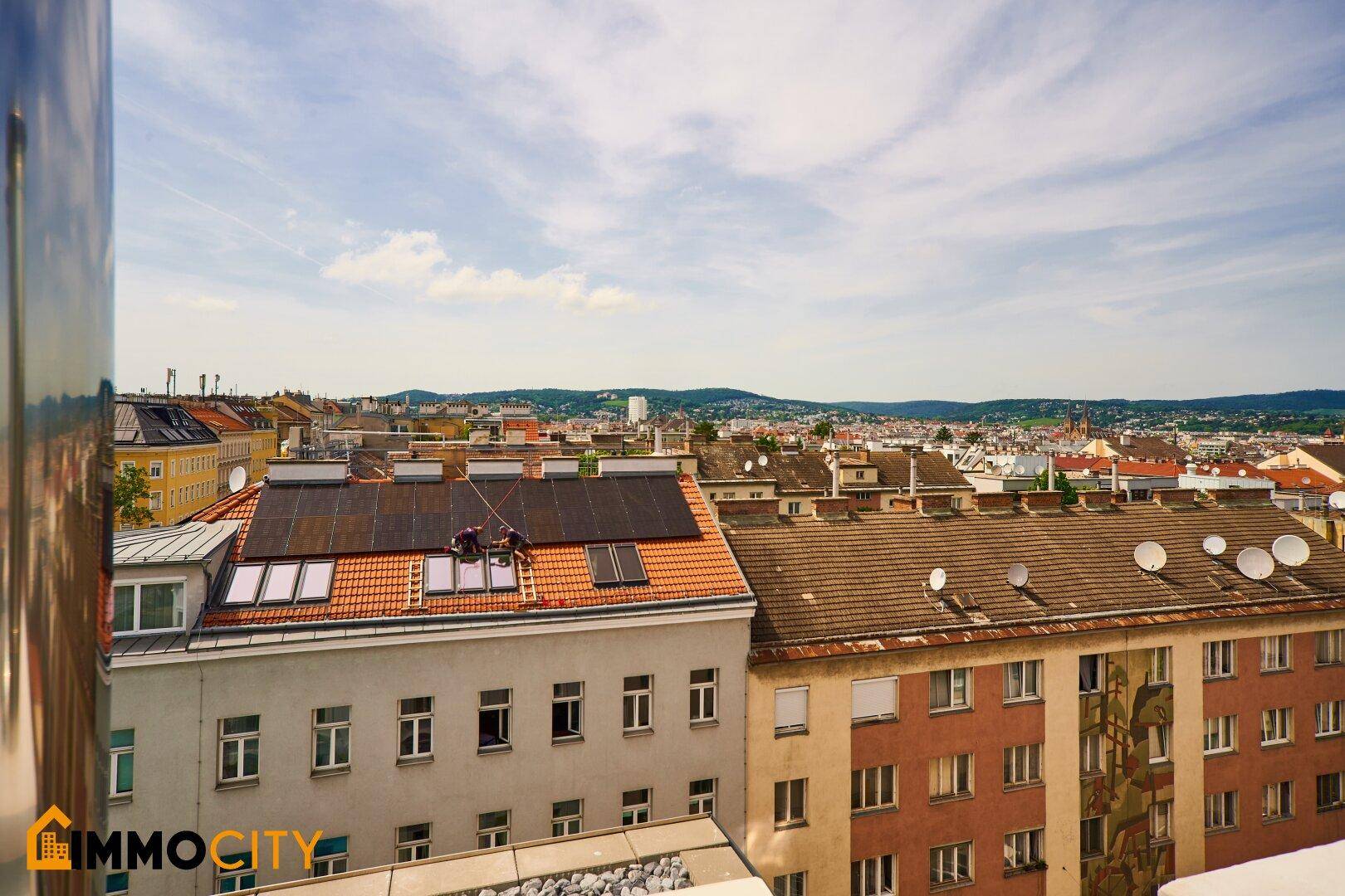 Blick von oben auf ein Dach mit Solarpaneelen und Dachfenstern, umgeben von Stadtgebäuden.