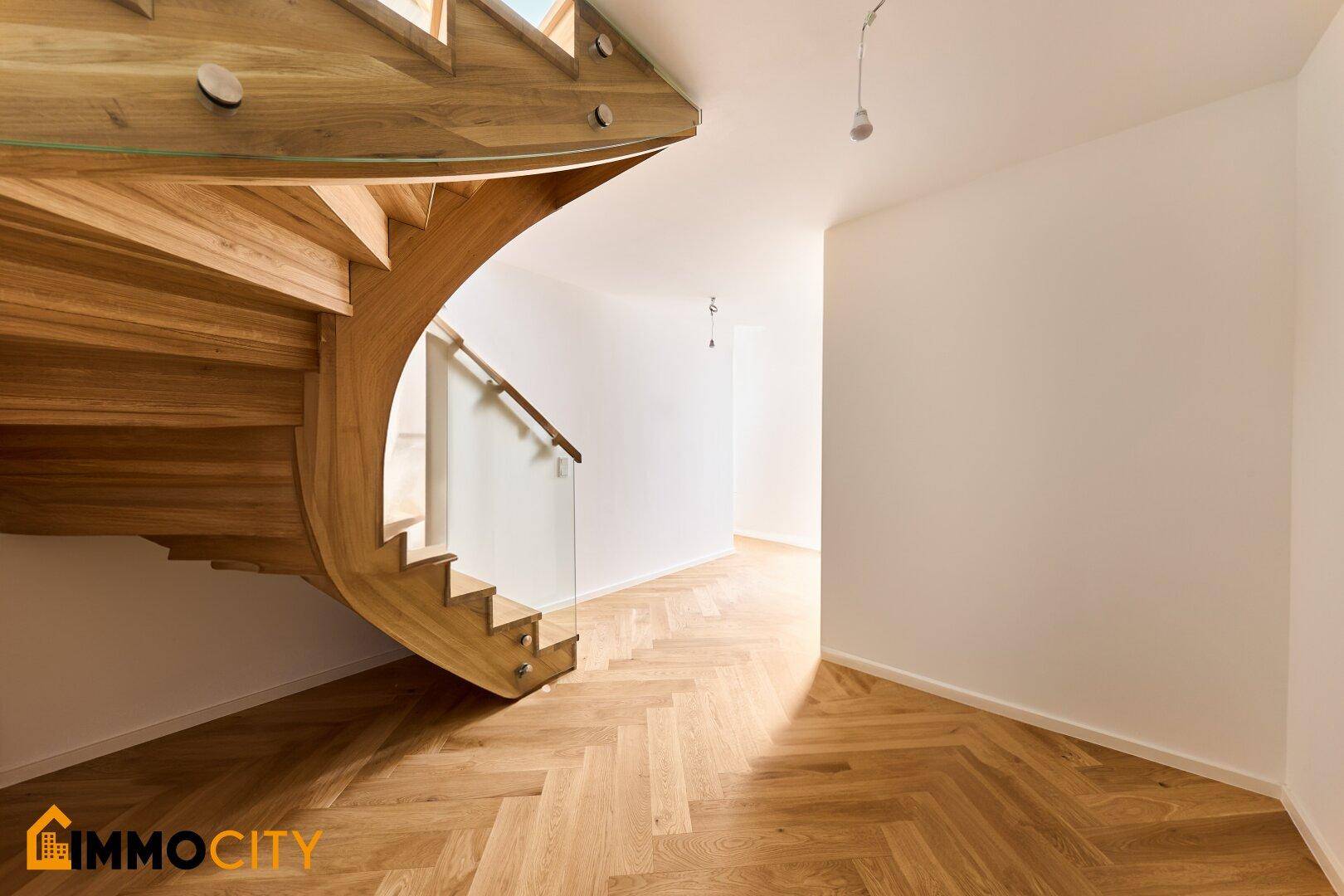 Elegante Holzwendeltreppe mit Glasgeländer und Fischgrätparkett in einem hellen, modernen Flur.