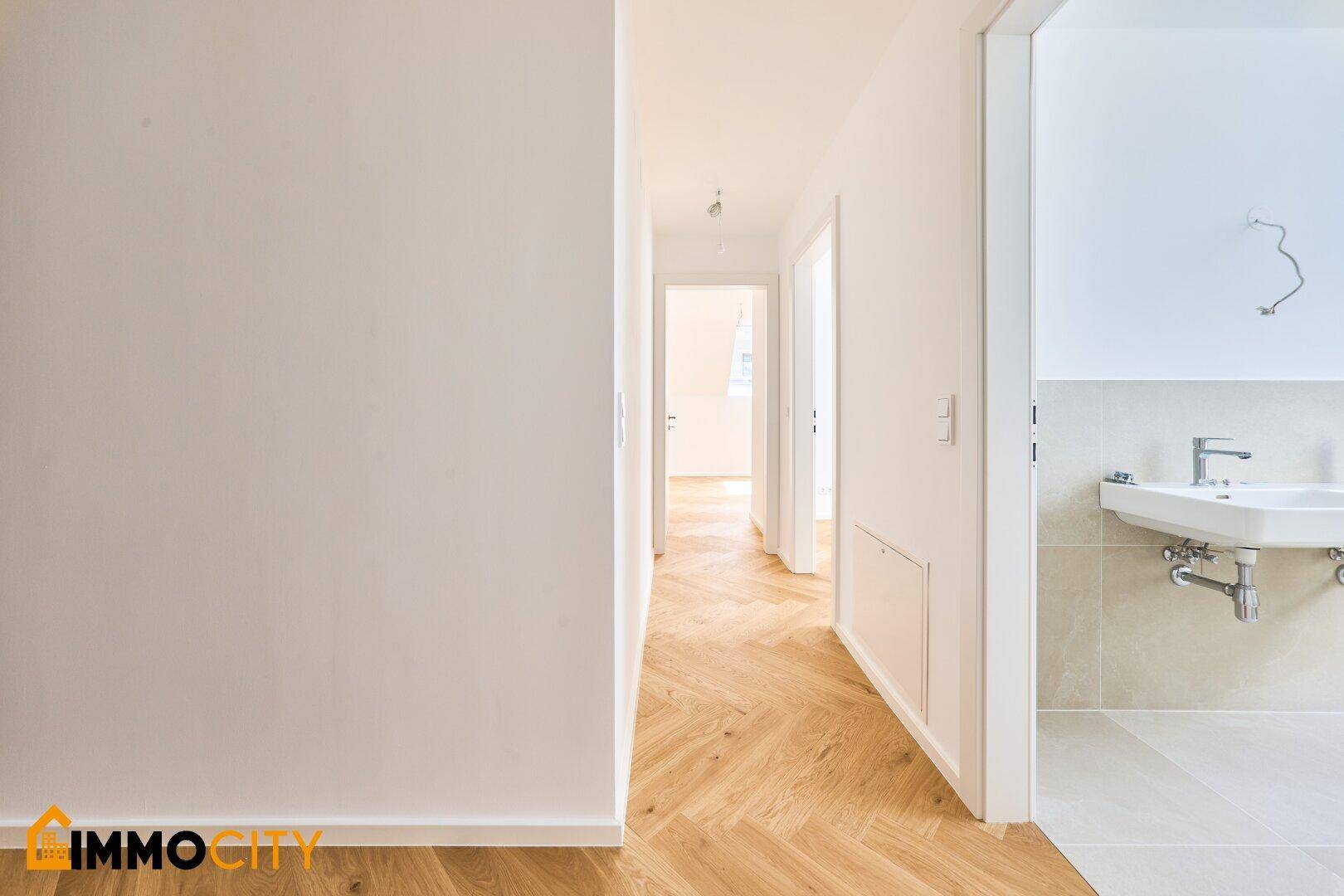 Langer, heller Flur mit Fischgrätparkett und Zugang zu einem modernen Badezimmer mit Waschbecken.