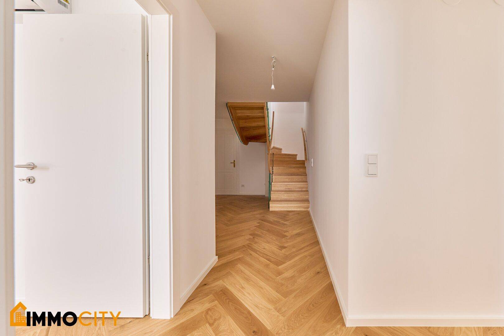 Heller Flur mit Fischgrätparkett und weißen Wänden, der zu einer eleganten Holztreppe führt.