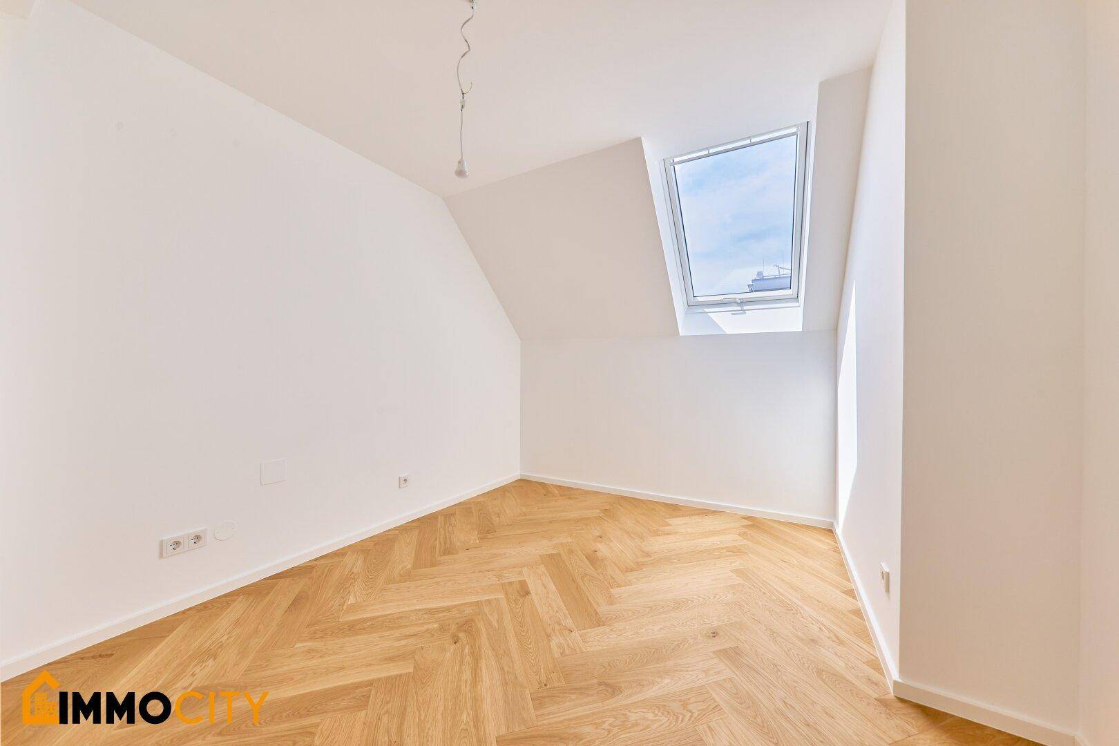 Helles Zimmer im Dachgeschoss mit Fischgrätparkett, weißen Wänden und einem großen Dachfenster.