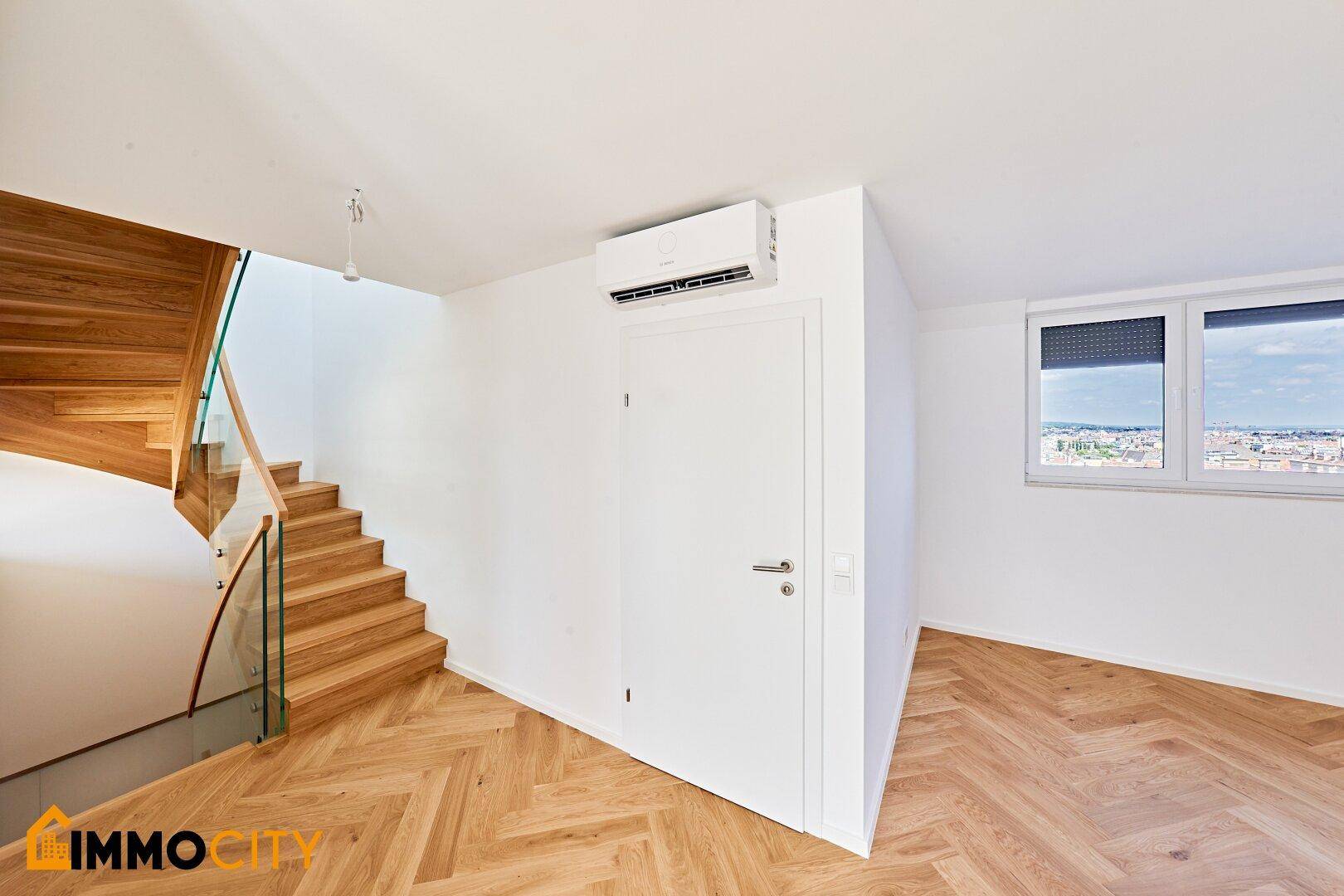 Heller Raum mit Fischgrätparkett, Holztreppe, Fenster mit Rollladen und Klimaanlage.