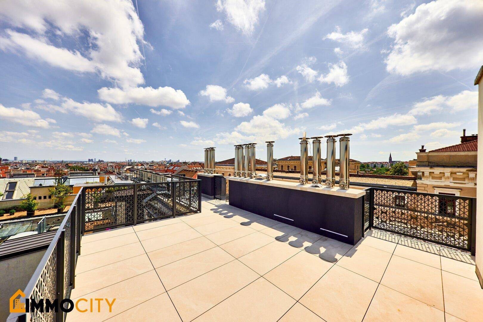 Geräumige Dachterrasse mit Stadtblick, Fliesenboden und modernen Schornsteinen unter blauem Himmel.