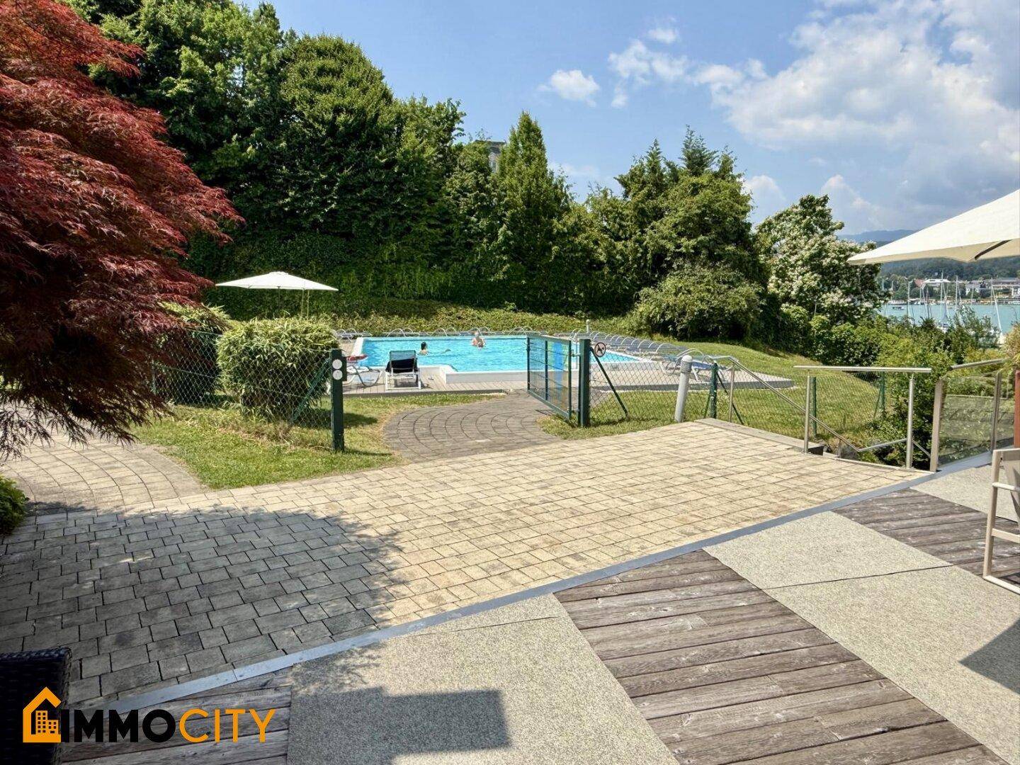 Außenpoolbereich mit Liegewiese, umgeben von Grünflächen und einem gepflasterten Terrassenbereich.