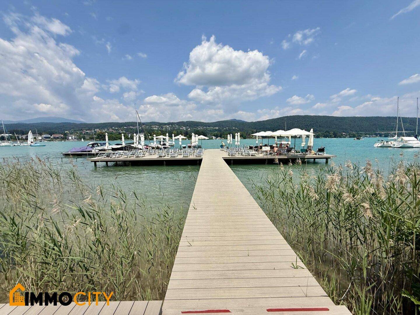 Holzsteg führt zu einer schwimmenden Plattform mit Sonnenliegen und Schirmen auf dem See.