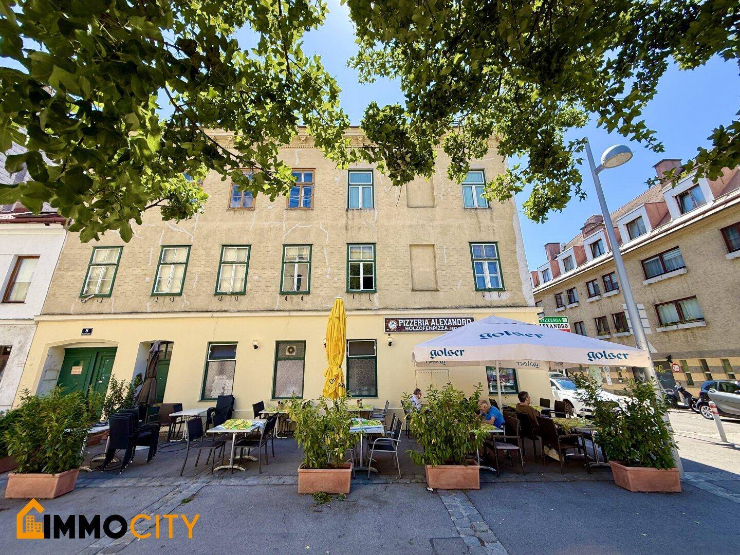 Außenansicht des Restaurants mit Terrasse, umgeben von grünen Bäumen unter blauem Himmel.