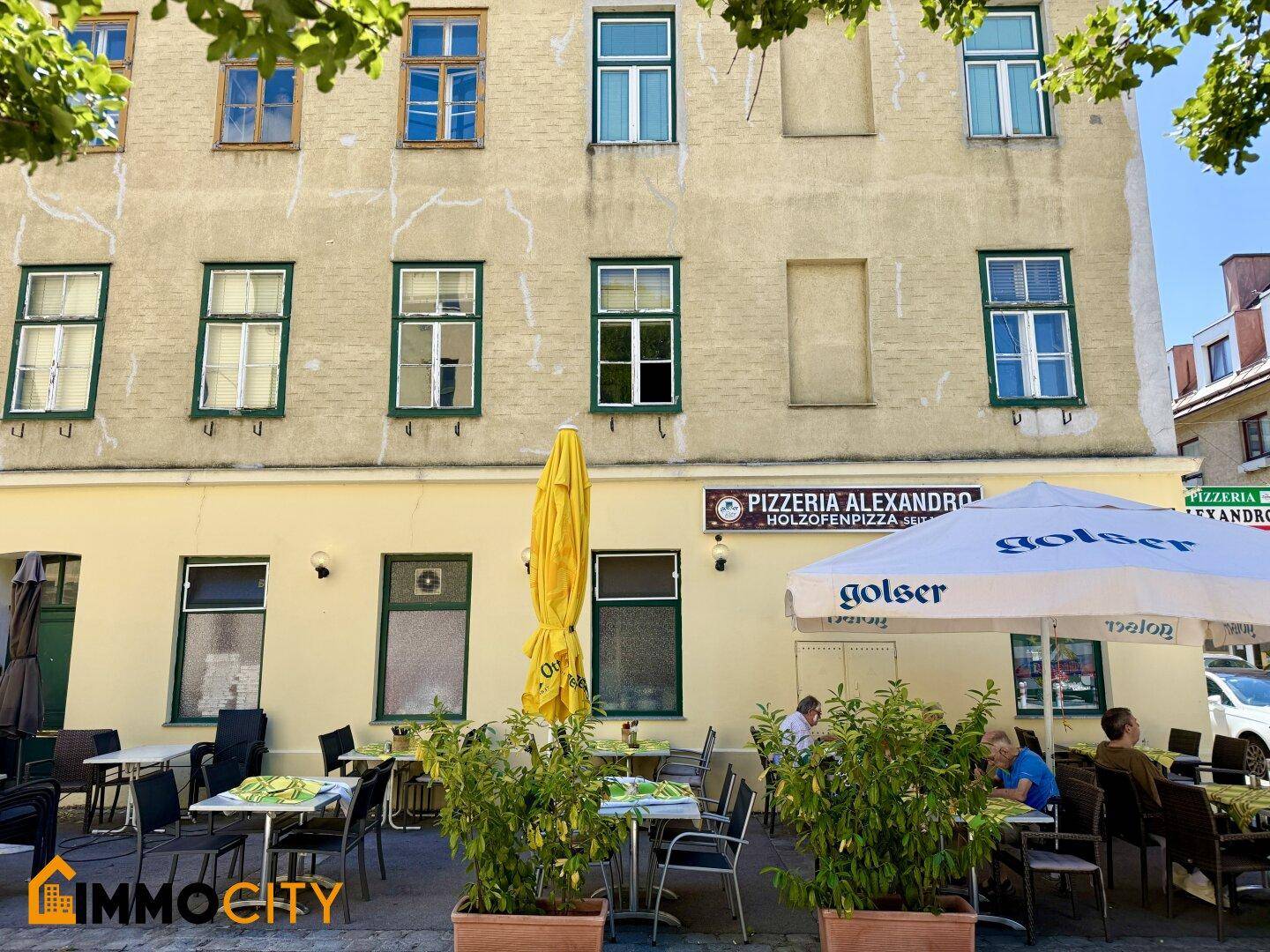 Frontansicht des Gebäudes mit der Restaurantterrasse und Sonnenschirmen unter strahlend blauem Himmel.