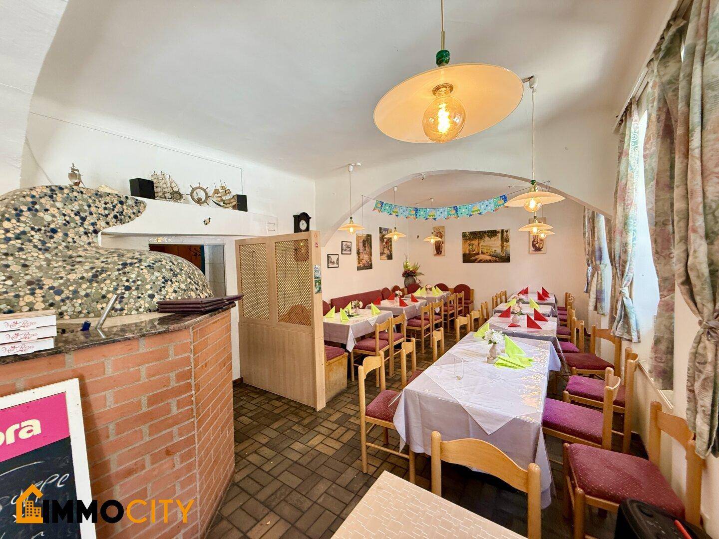 Gemütlicher Restaurant-Essbereich mit mehreren Tischen, Stühlen und einem traditionellen Pizzaofen im Hintergrund.