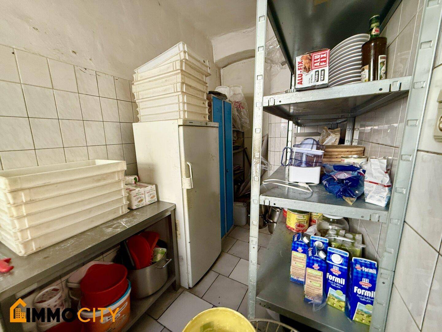 Lagerbereich einer Restaurantküche mit Regalen, einem Kühlschrank und gestapelten Pizzakartons.